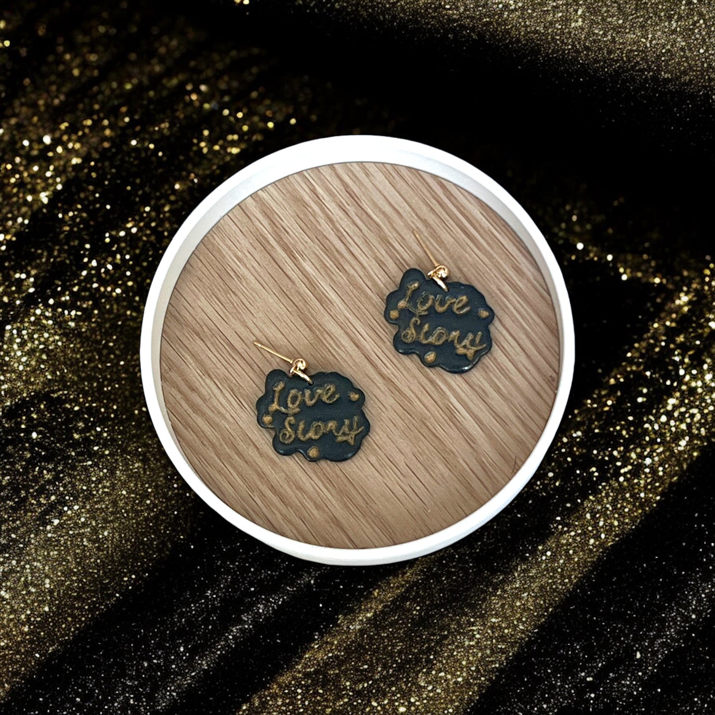Handmade "Love Story" earrings with gold text, resting on a wooden display dish and surrounded by shimmering black and gold fabric.