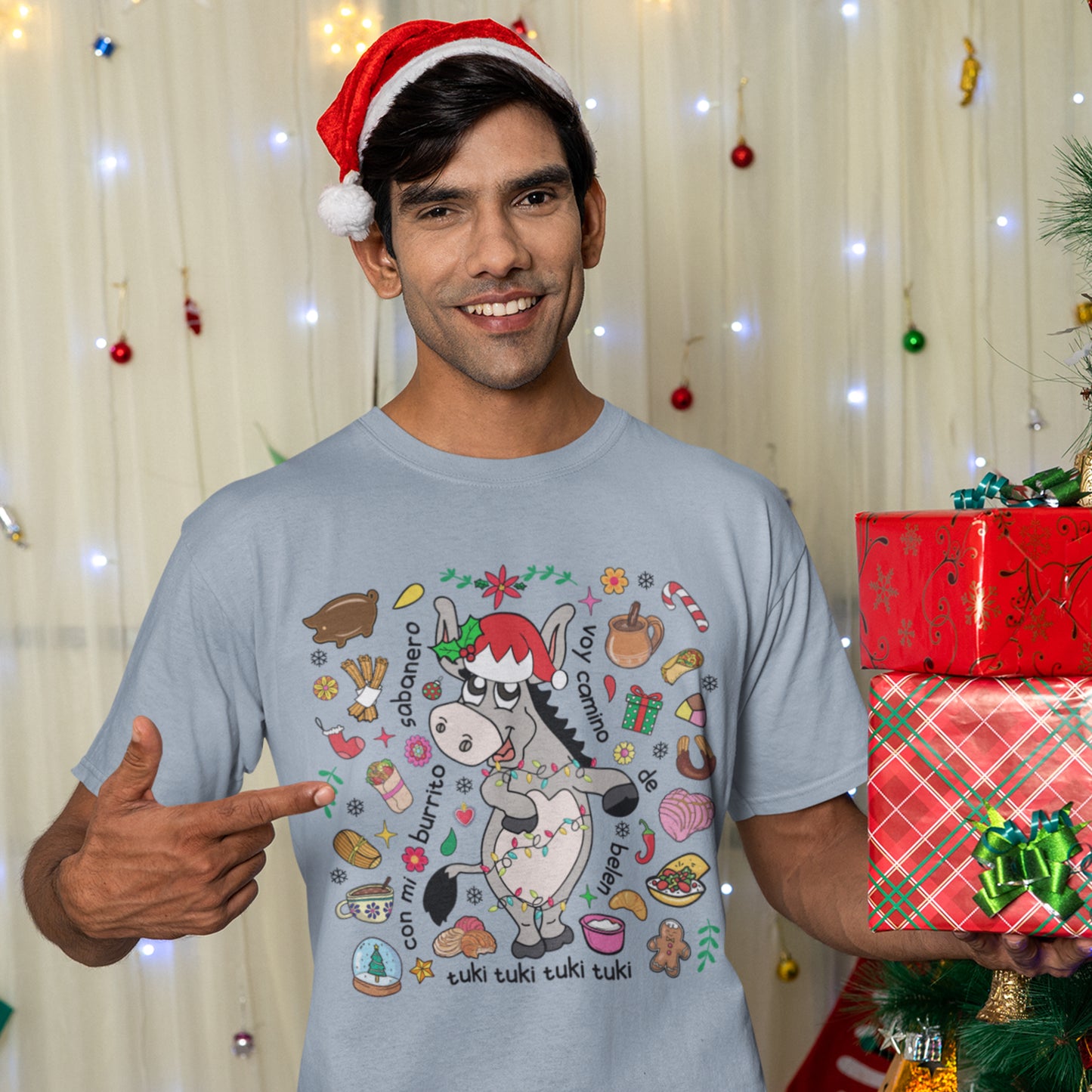 Man wearing a light blue "Burrito Sabanero" t-shirt with colorful holiday graphics and lyrics, posing with gifts and festive decor.