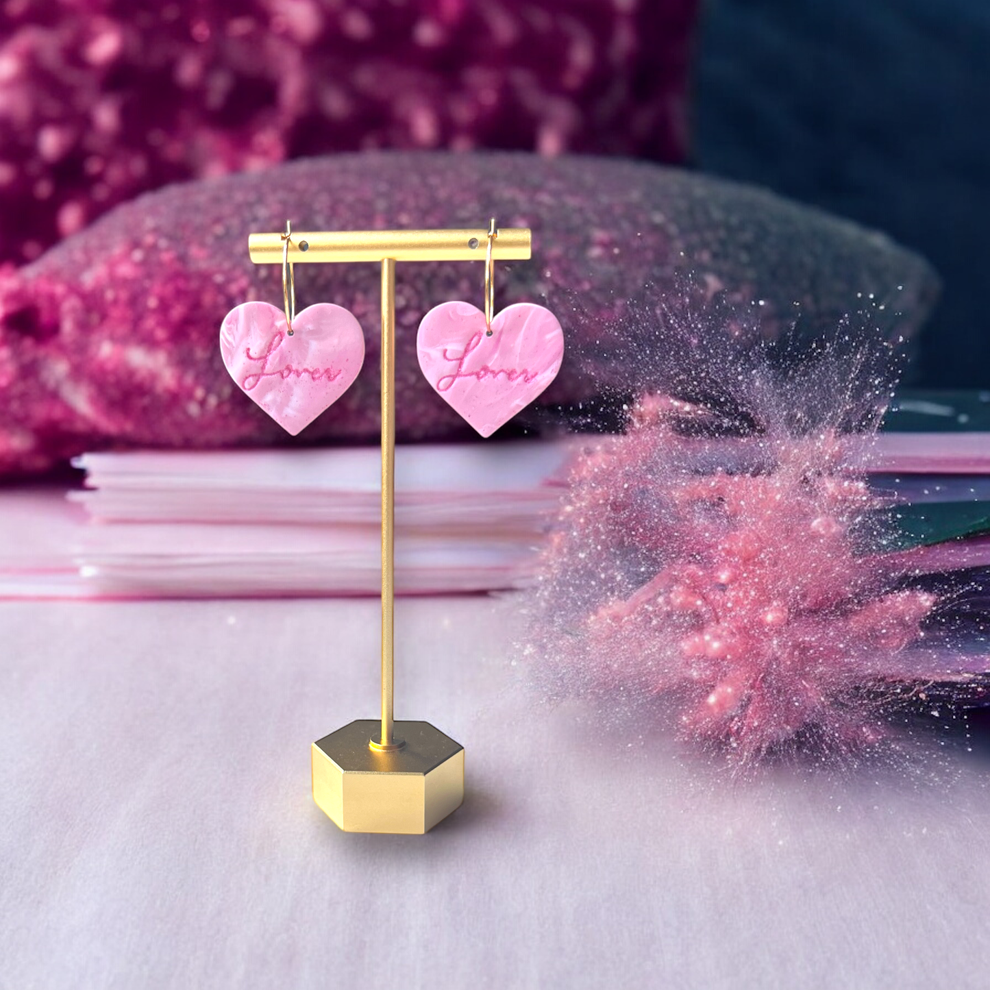 Elegant marbled pink heart-shaped drop earrings with "Lover" engraving, hanging from a gold stand with a dreamy, soft pink cosmic backdrop.