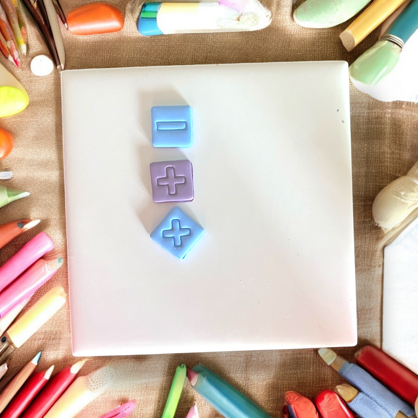 Three stamped clay math symbols neatly aligned on a white tile, surrounded by colorful school supplies.