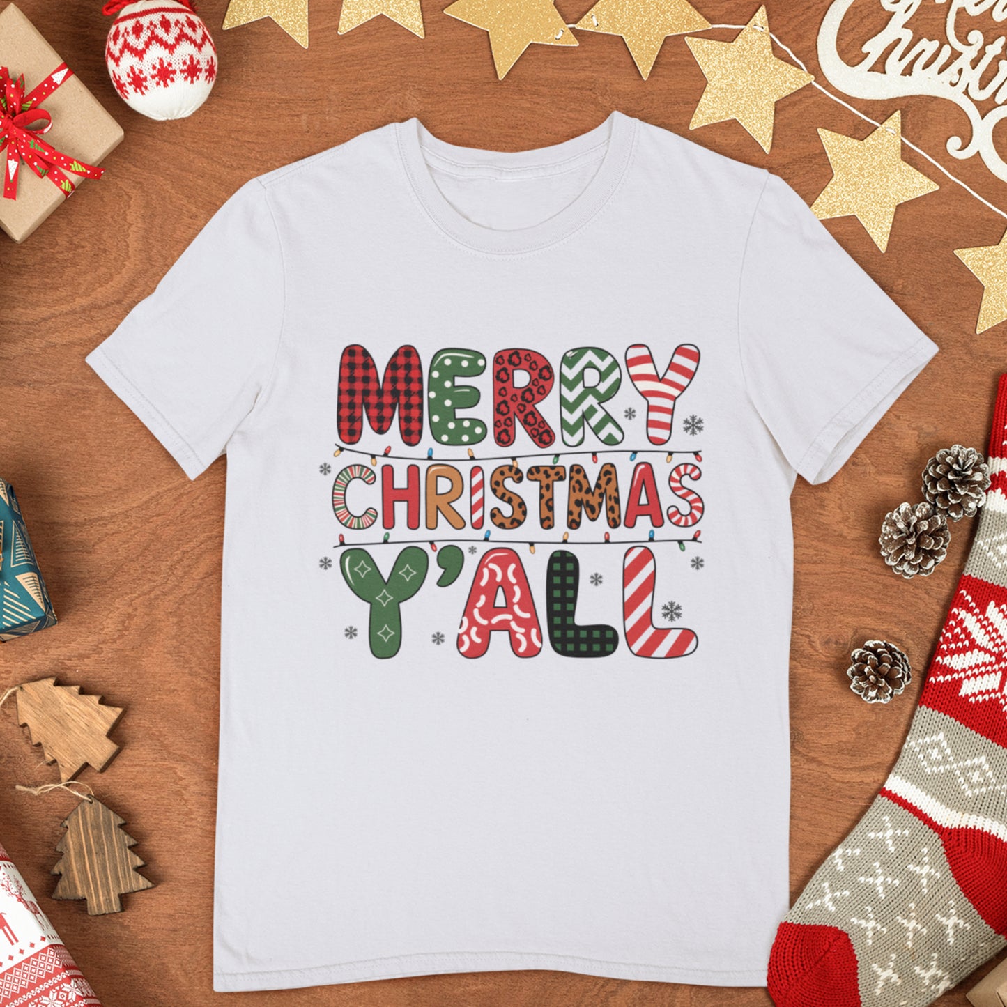 Flat lay of a white "Merry Christmas Y'all" t-shirt with colorful festive text, displayed on a wooden surface with holiday decor.