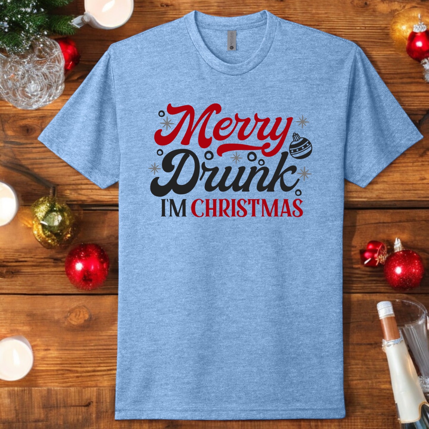 Flat lay of a light blue "Merry Drunk I’m Christmas" t-shirt with festive red and black text, displayed with holiday-themed decor.