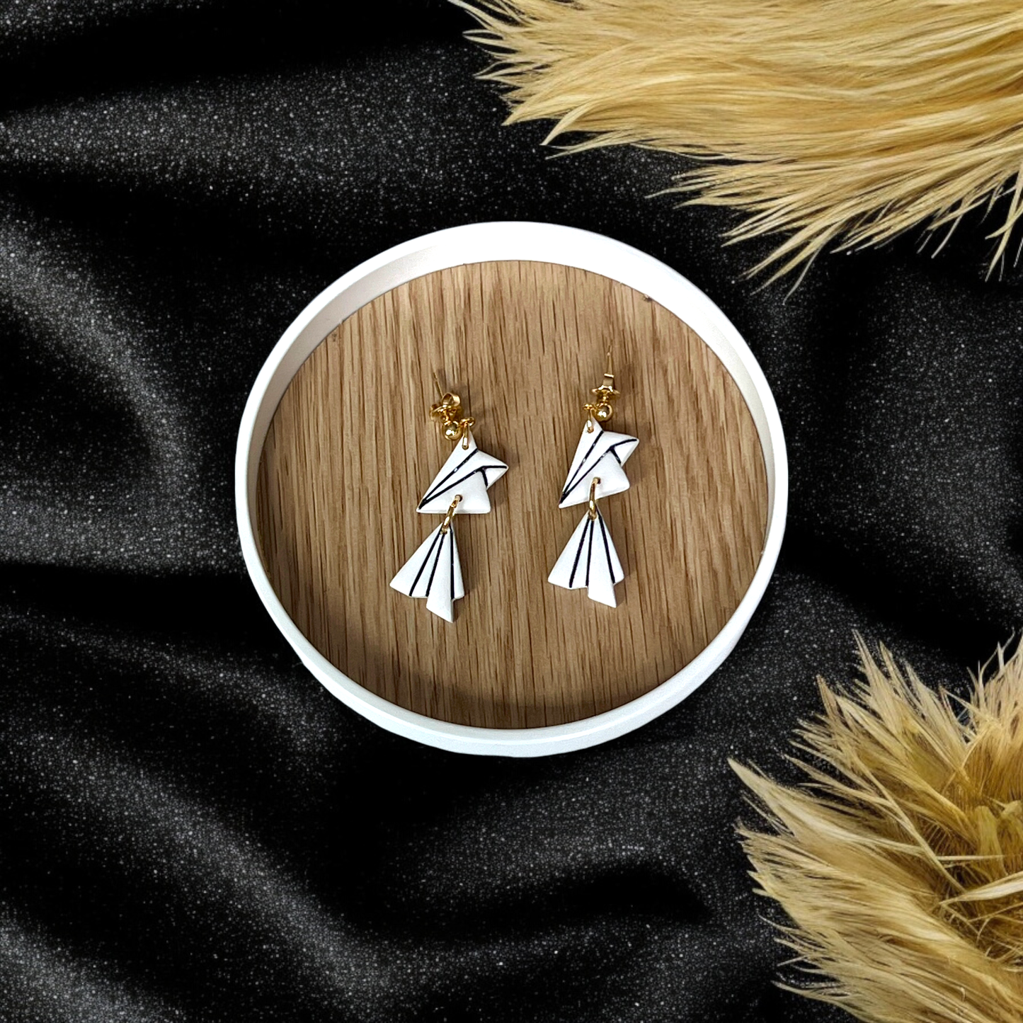 Elegant white drop earrings featuring black linear accents and a layered geometric shape, displayed on a wooden tray with a black fabric background.