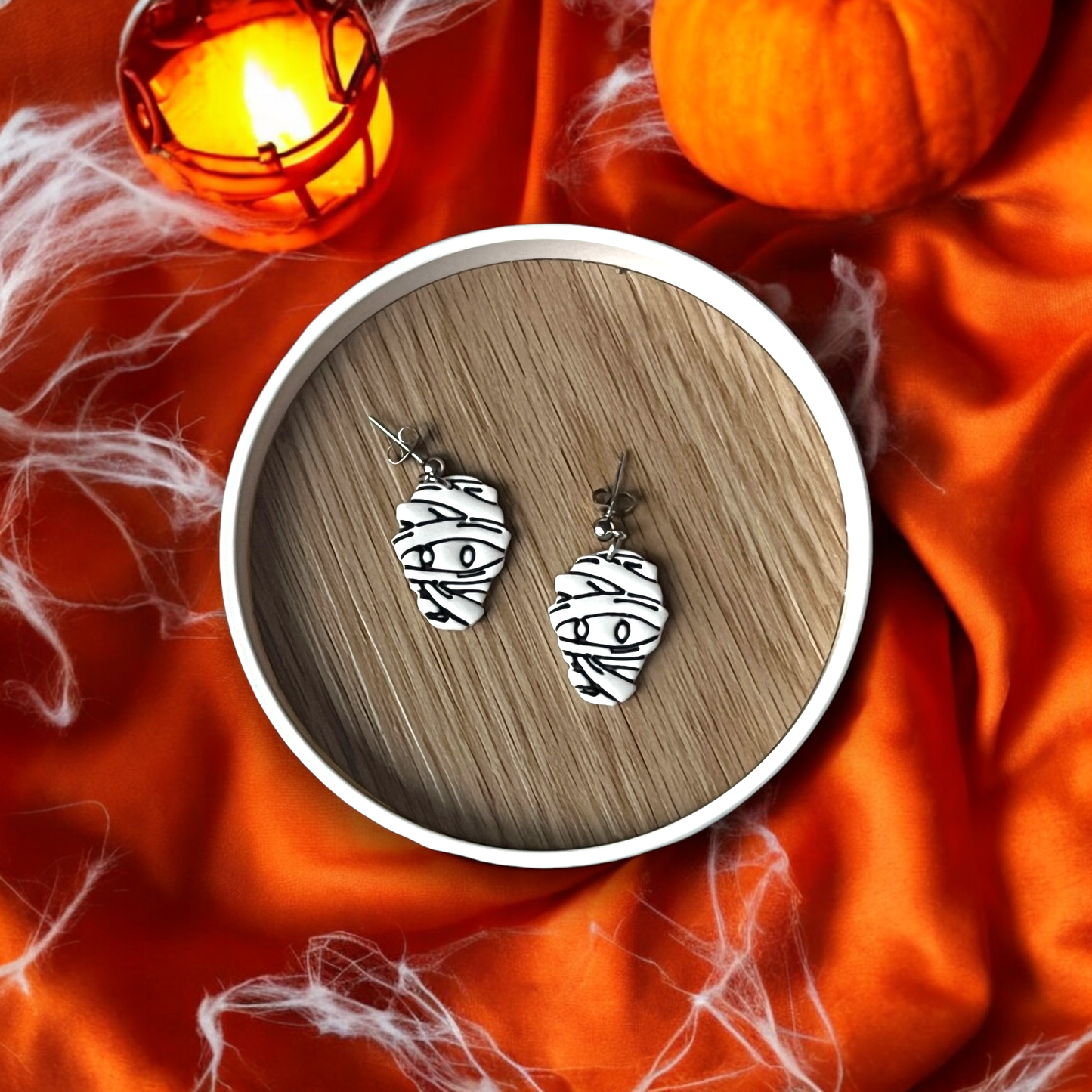 Pair of Halloween-themed mummy face earrings featuring black bandage lines, placed on a wooden tray surrounded by cobwebs and pumpkin decor.