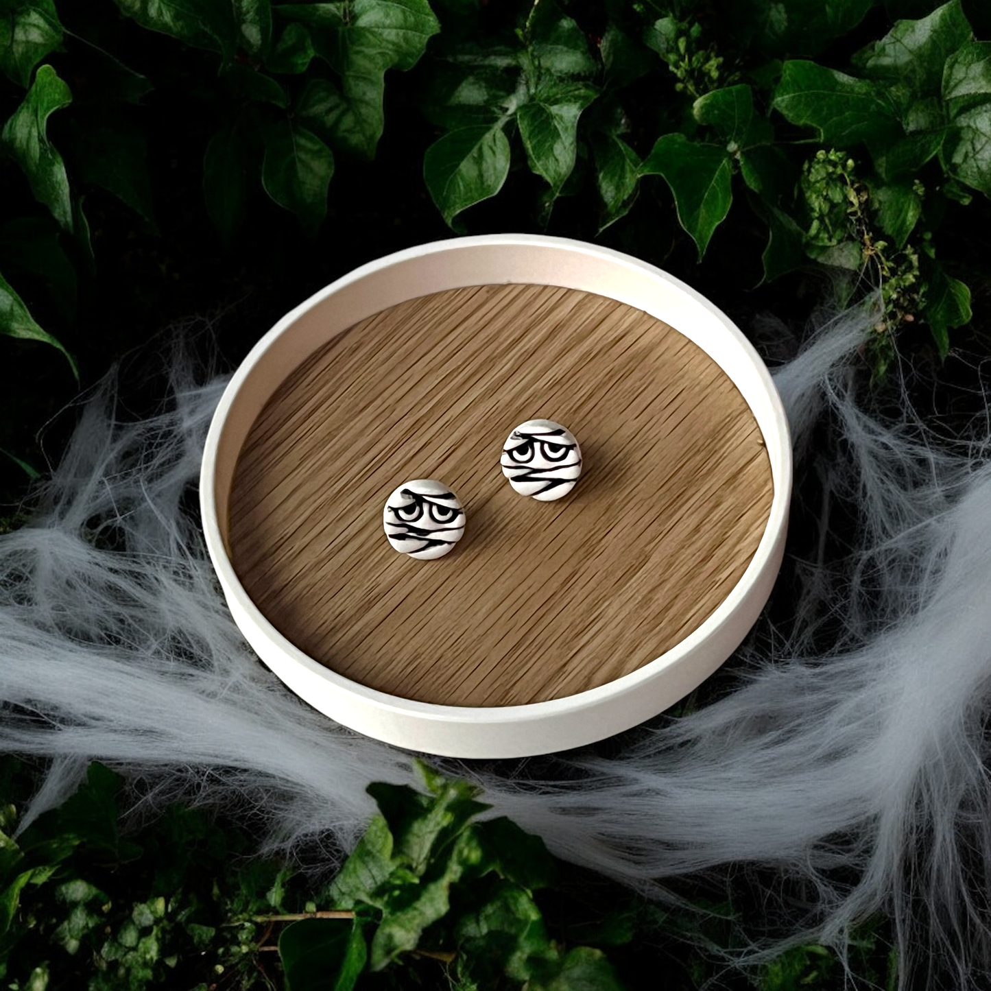 Round black-and-white mummy face stud earrings with spooky bandage design, displayed on a branded card surrounded by cobwebs and ivy.