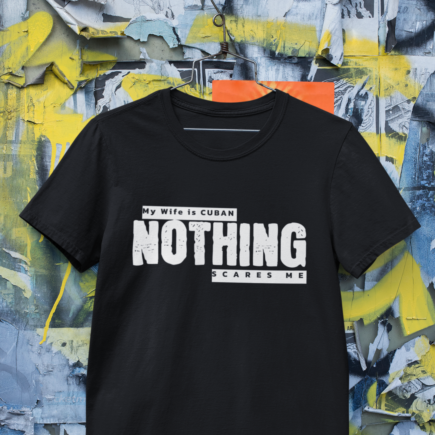 Black "My Wife is Cuban, Nothing Scares Me" t-shirt hanging on a hanger against an artistic, industrial background.