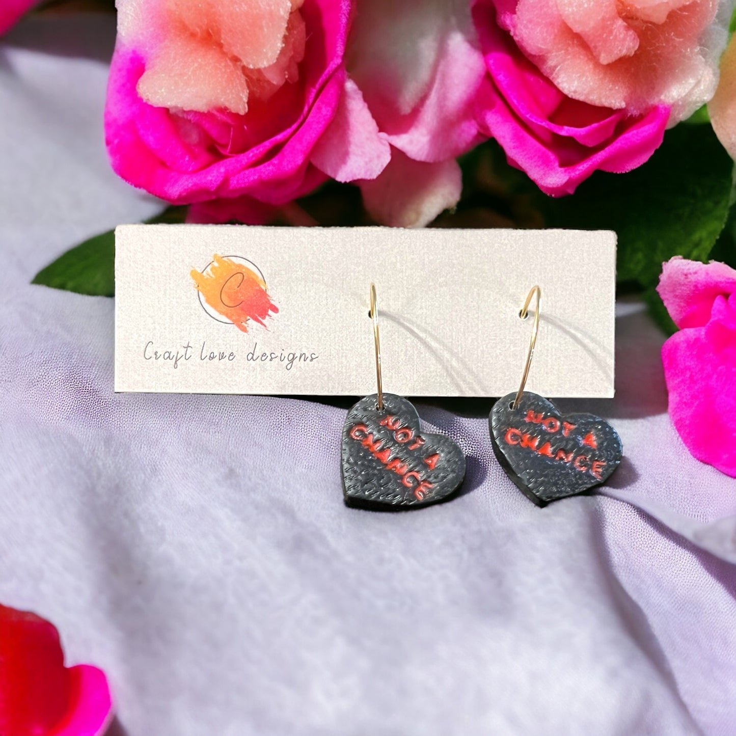 Black heart-shaped hoop dangle earrings with “Not A Chance” text in red displayed on a white card, surrounded by pink roses.