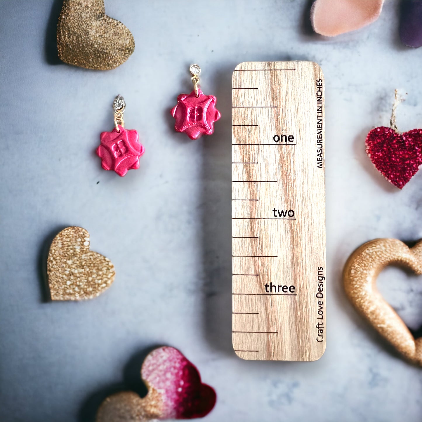 A pair of red cubic zirconia dainty dangle earrings displayed beside a ruler for size reference, surrounded by glittery hearts and decorative props.