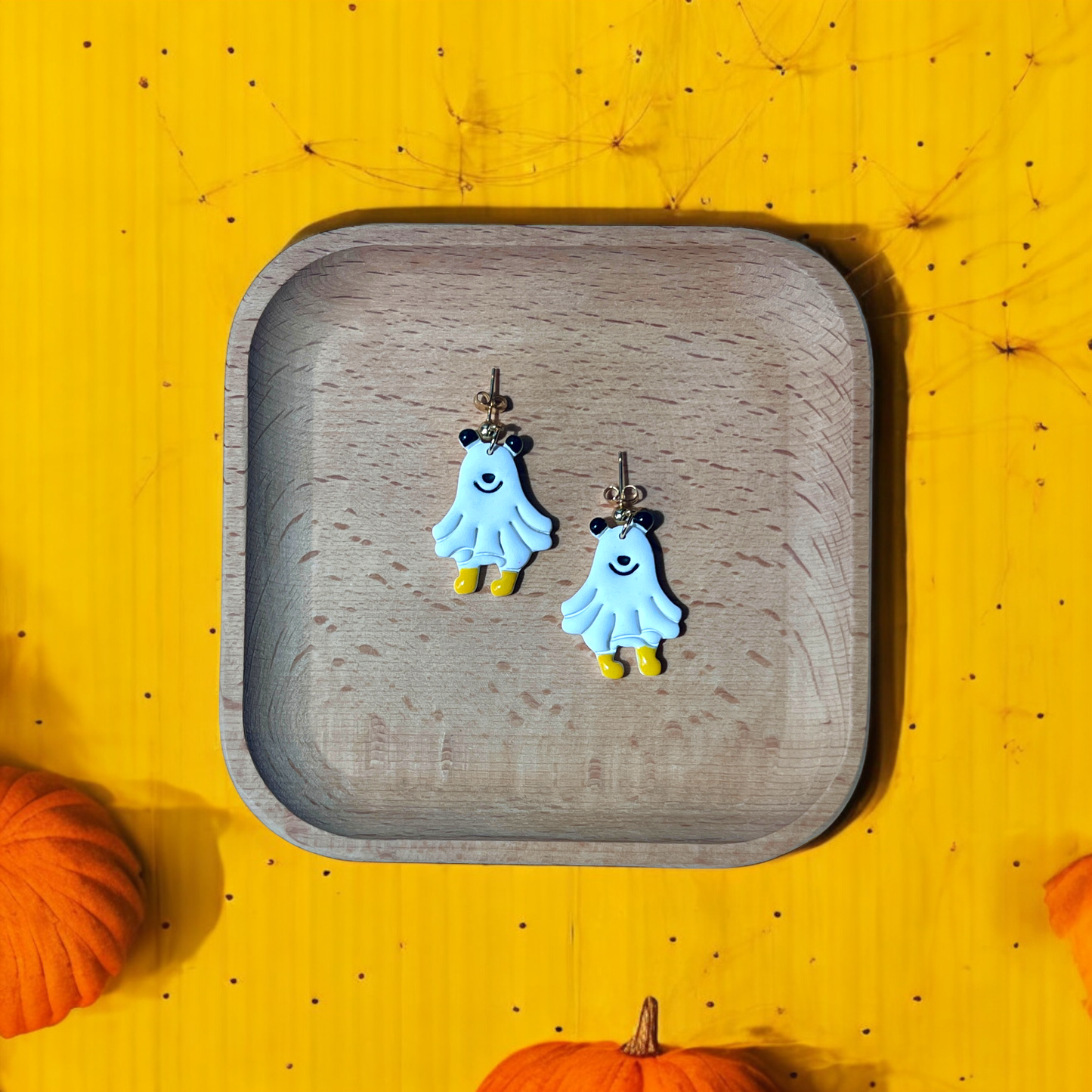 Adorable ghost earrings with yellow boots displayed on a wooden plate, surrounded by pumpkins and a vibrant yellow background.
