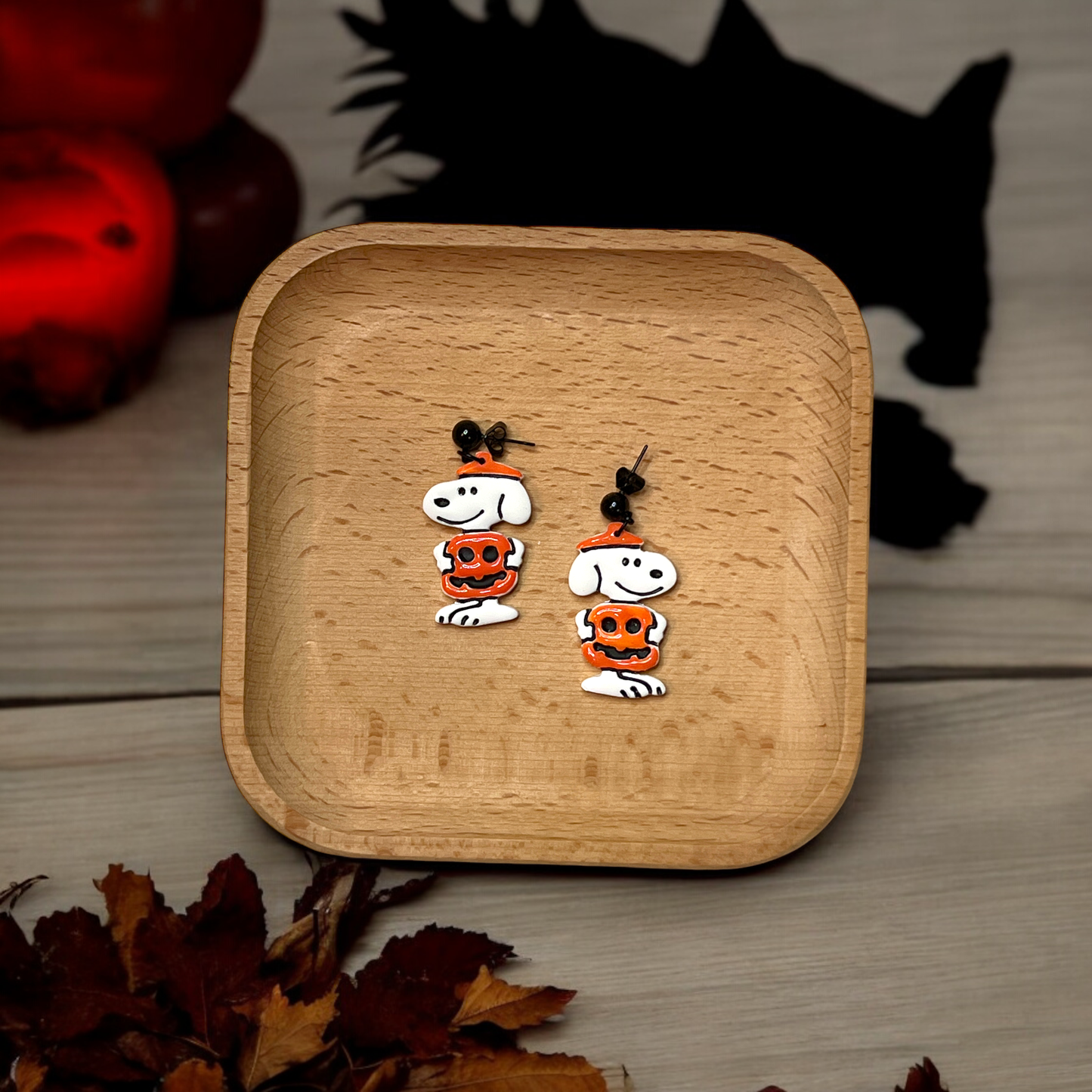 White polymer clay dog earrings with an orange pumpkin design displayed on a wooden tray, surrounded by a spooky Halloween-themed setting.