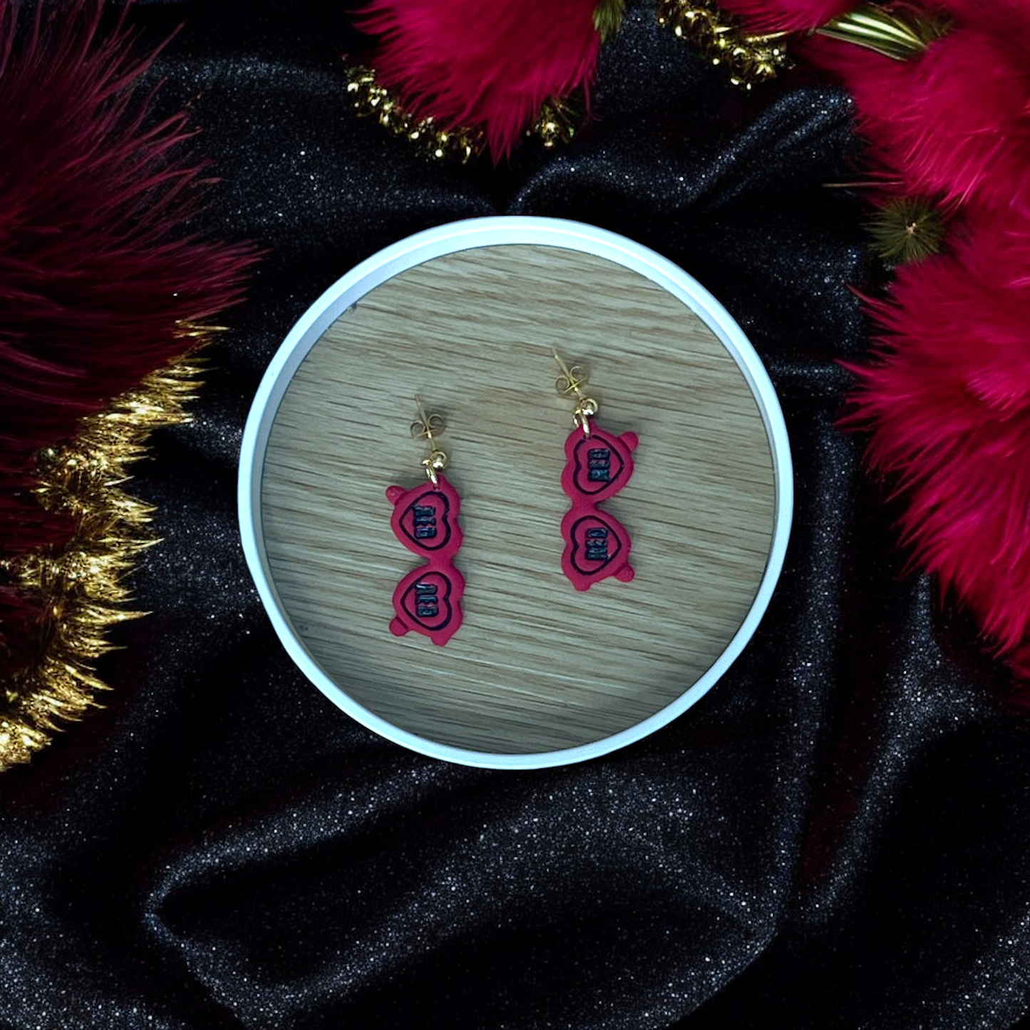 Red heart sunglasses-shaped drop earrings placed in a wooden tray, surrounded by festive black velvet, gold accents, and red feathers.