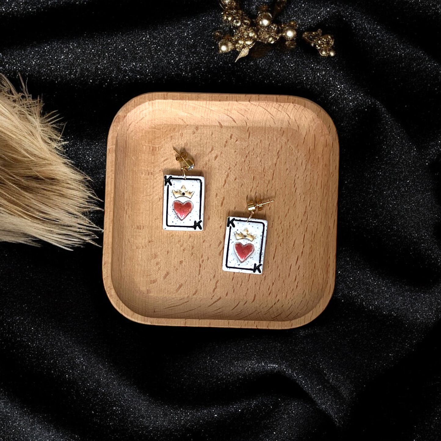 Pair of King of Hearts-themed earrings showcasing a crowned red heart on a white card design, displayed in a wooden tray against black fabric.