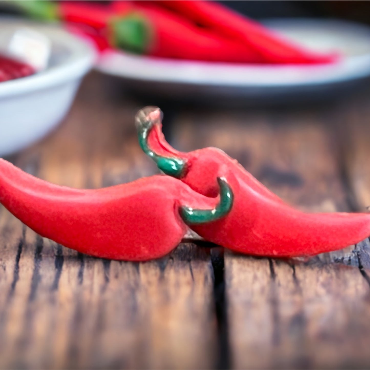 A detailed close-up of Red Hot Chili Pepper stud earrings, showcasing their curved red shape and glossy green tops on a wooden surface.