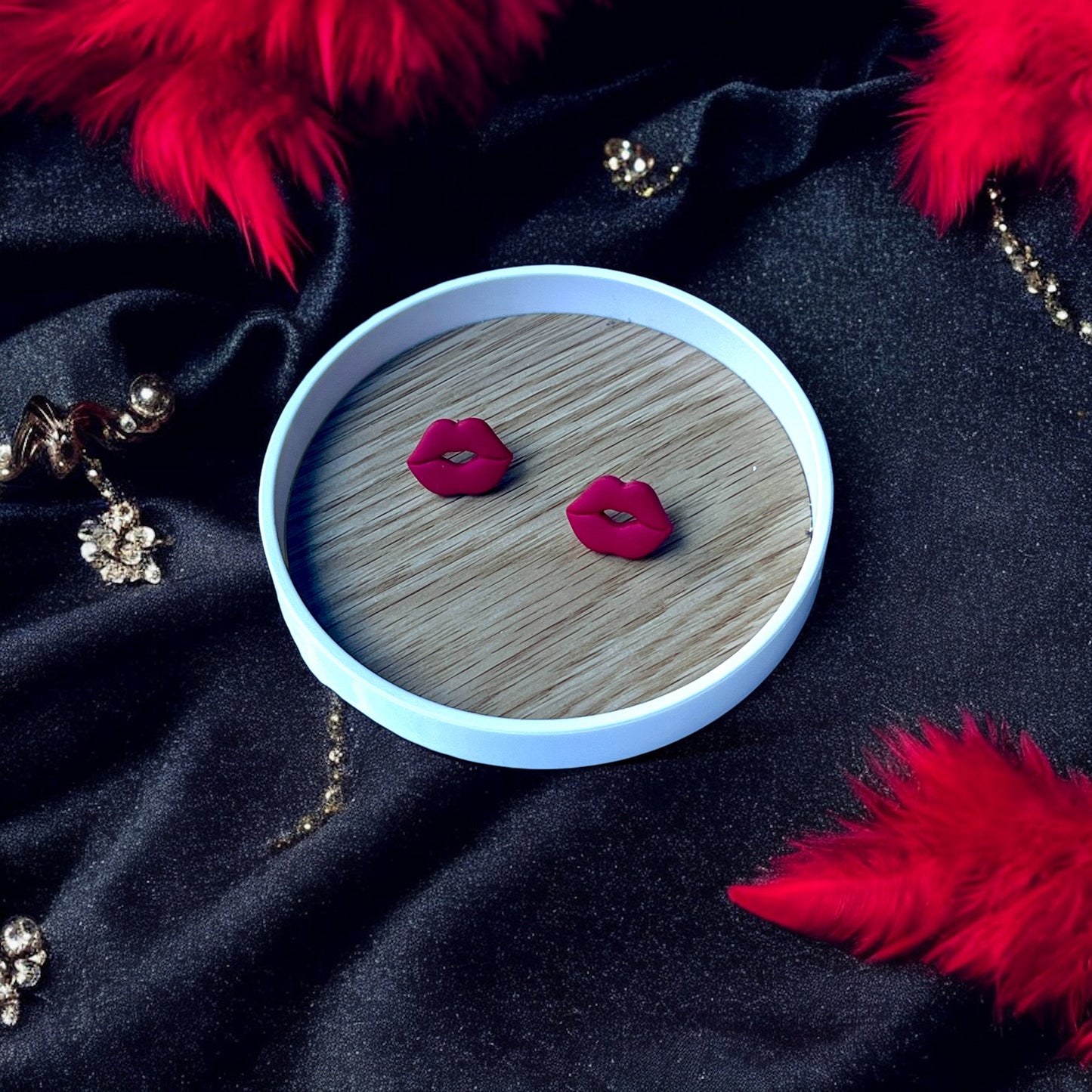 Red lips stud earrings placed on a wooden tray, surrounded by luxurious black fabric, sparkling embellishments, and vibrant red feathers.