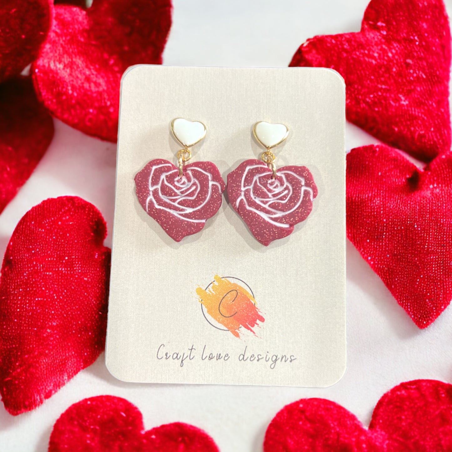 Elegant red rose dangle earrings with glitter and white heart studs displayed on a branded card surrounded by plush red hearts.