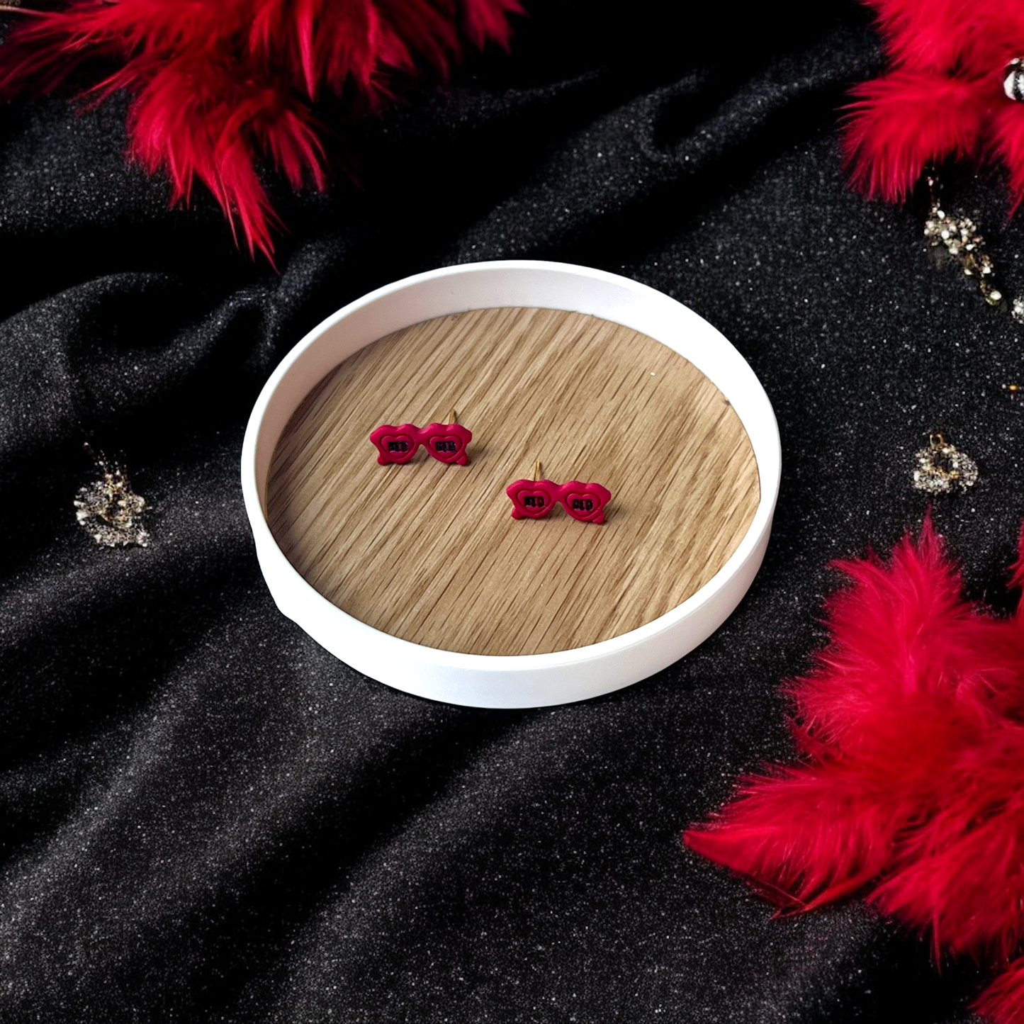 Retro red sunglasses earrings with "Red" text placed in a wooden tray, surrounded by red feathers and black sparkly fabric background.
