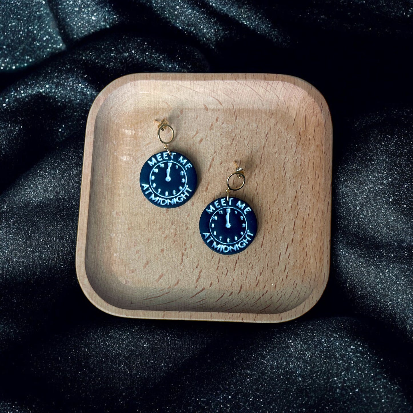 Round black earrings with midnight clock detailing and "Meet Me At Midnight" text, resting in a wooden tray on sparkling black fabric.