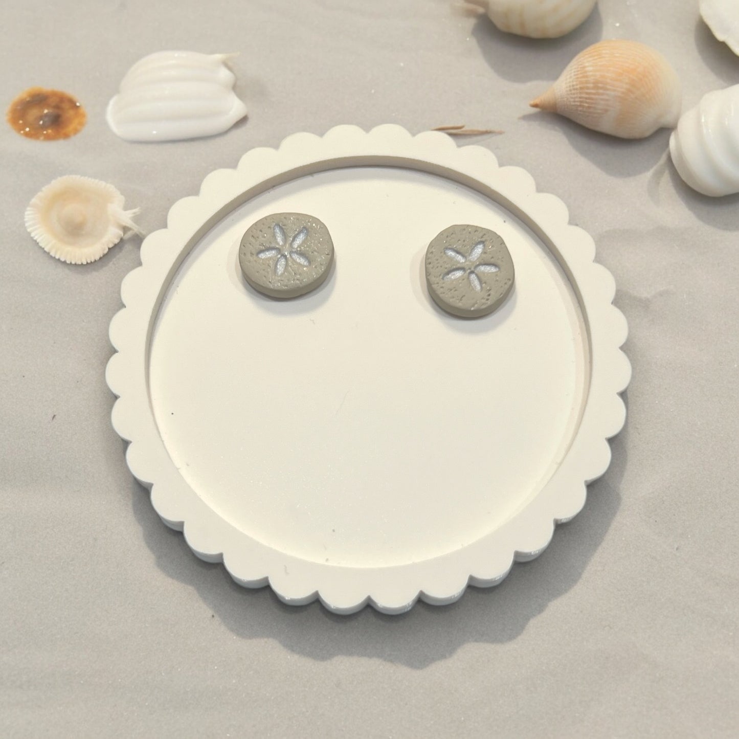 Clay sand dollar earrings placed on a scalloped white tray, surrounded by delicate seashells, creating a soft and beachy aesthetic.
