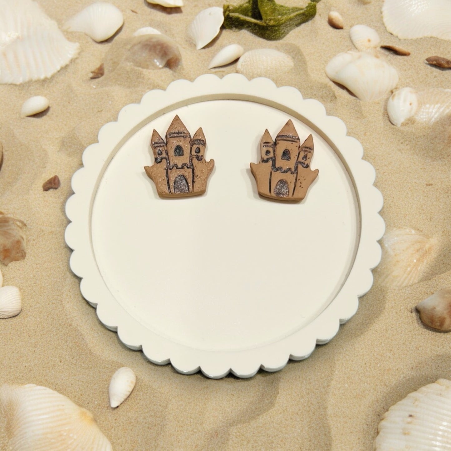 Beach-inspired sandcastle earrings displayed on a round white tray, surrounded by seashells on sandy beach decor.