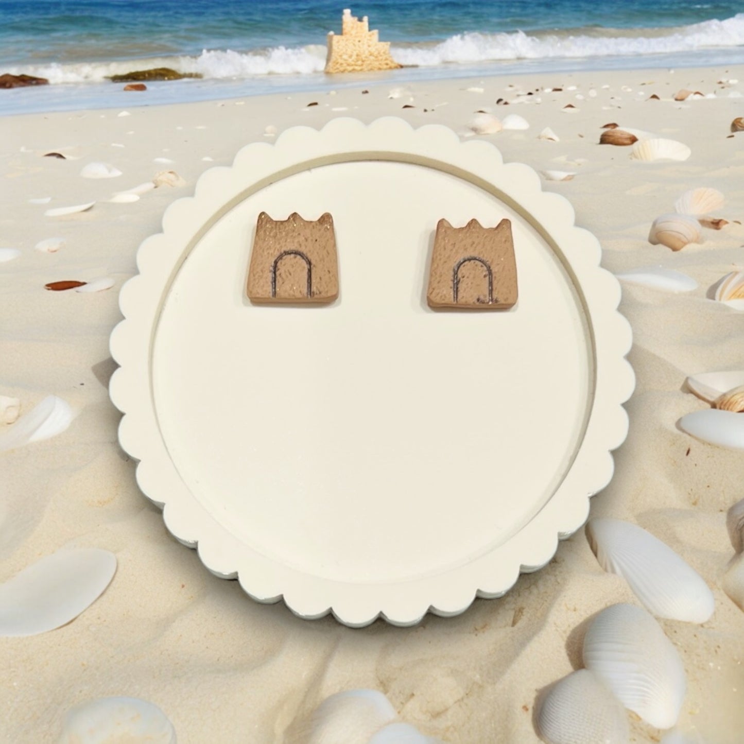Sandcastle earrings resting on a decorative round white tray, surrounded by seashells and sand.