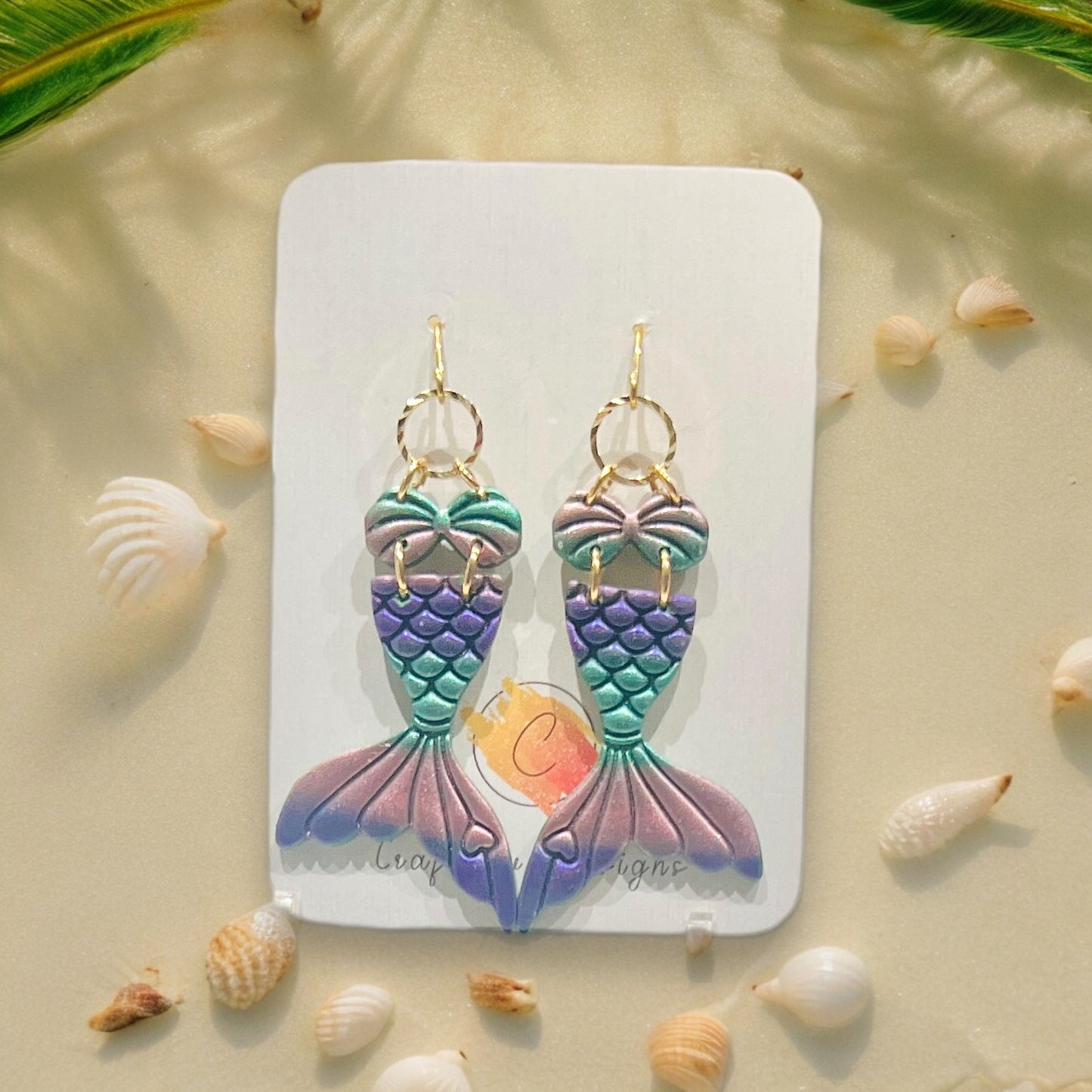 Multicolored mermaid tail earrings with a shiny finish displayed on a jewelry card, surrounded by tropical leaves and seashells for a beachy touch.
