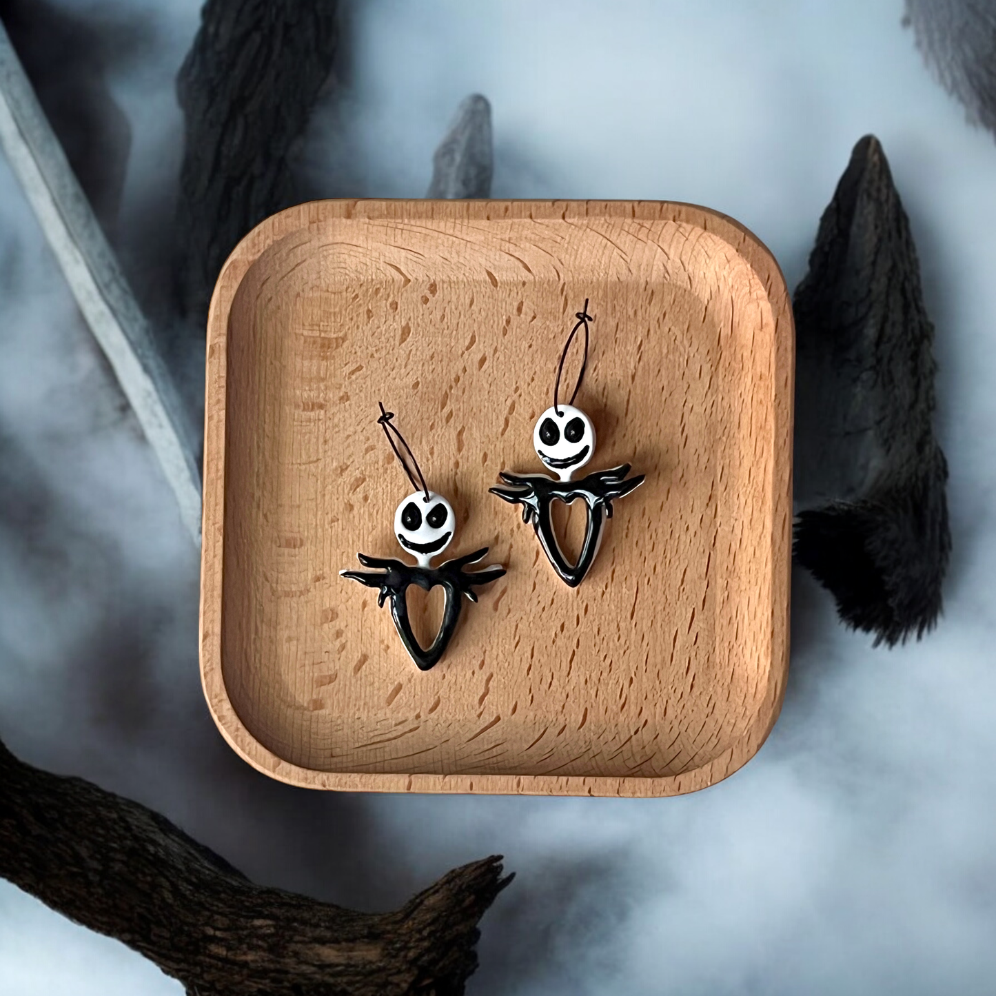 Pair of spooky skeleton dangle earrings with heart-shaped cutouts, placed on a wooden tray against a foggy Halloween backdrop.