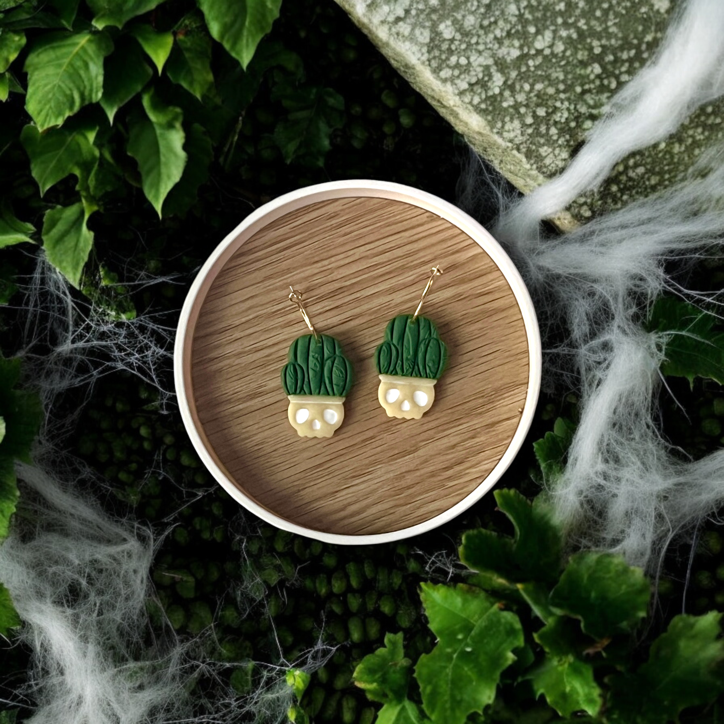 Unique skull planter earrings with textured green leaf tops, resting on a wooden tray amidst spooky cobweb decor and vibrant green foliage.