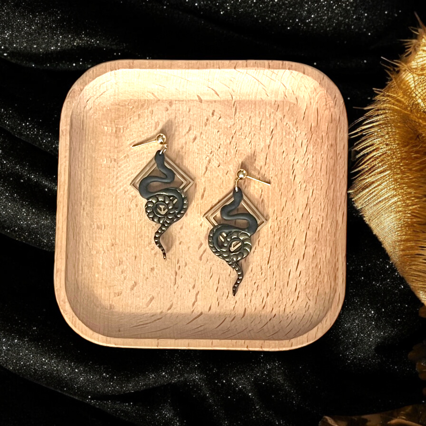 Black snake-shaped dangle earrings wrapped around diamond-shaped gold frames, presented on a wooden tray with golden and black decor accents.