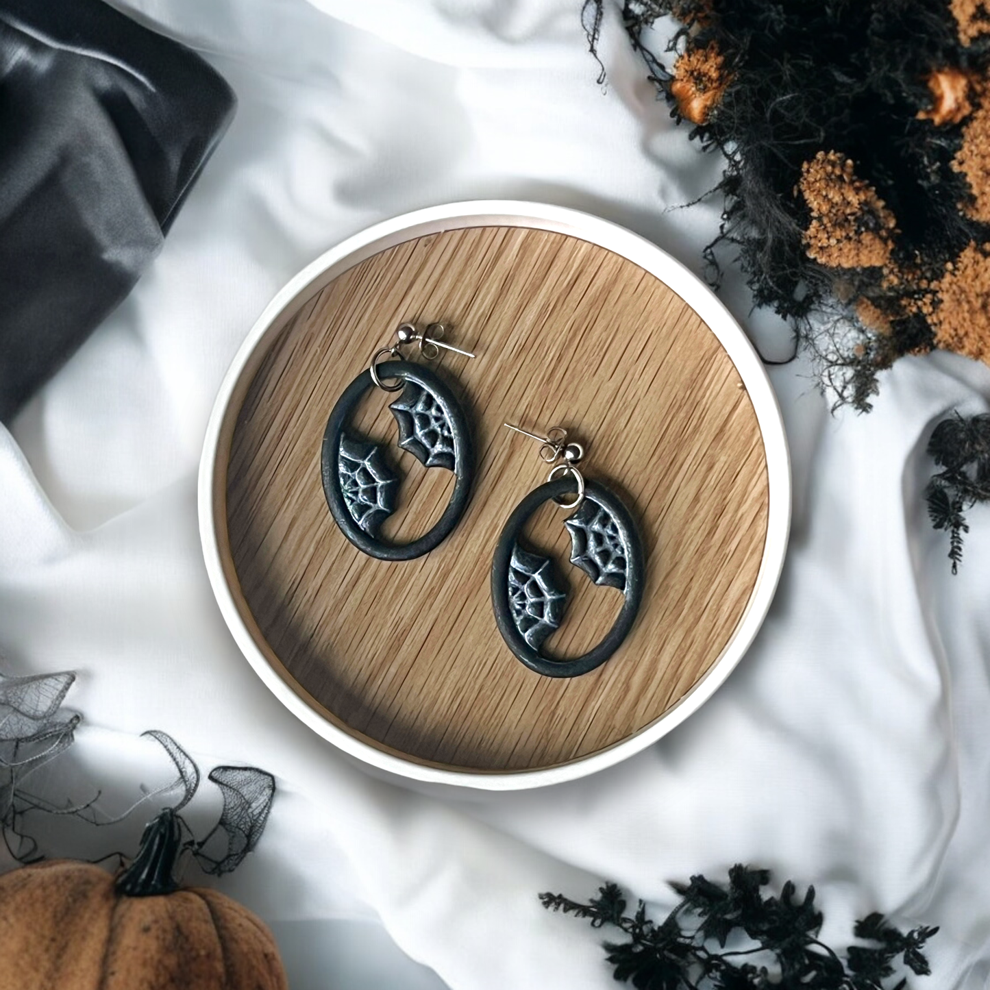 Oval-shaped earrings with spider web designs resting on a circular wooden tray, surrounded by white fabric and spooky seasonal accents.