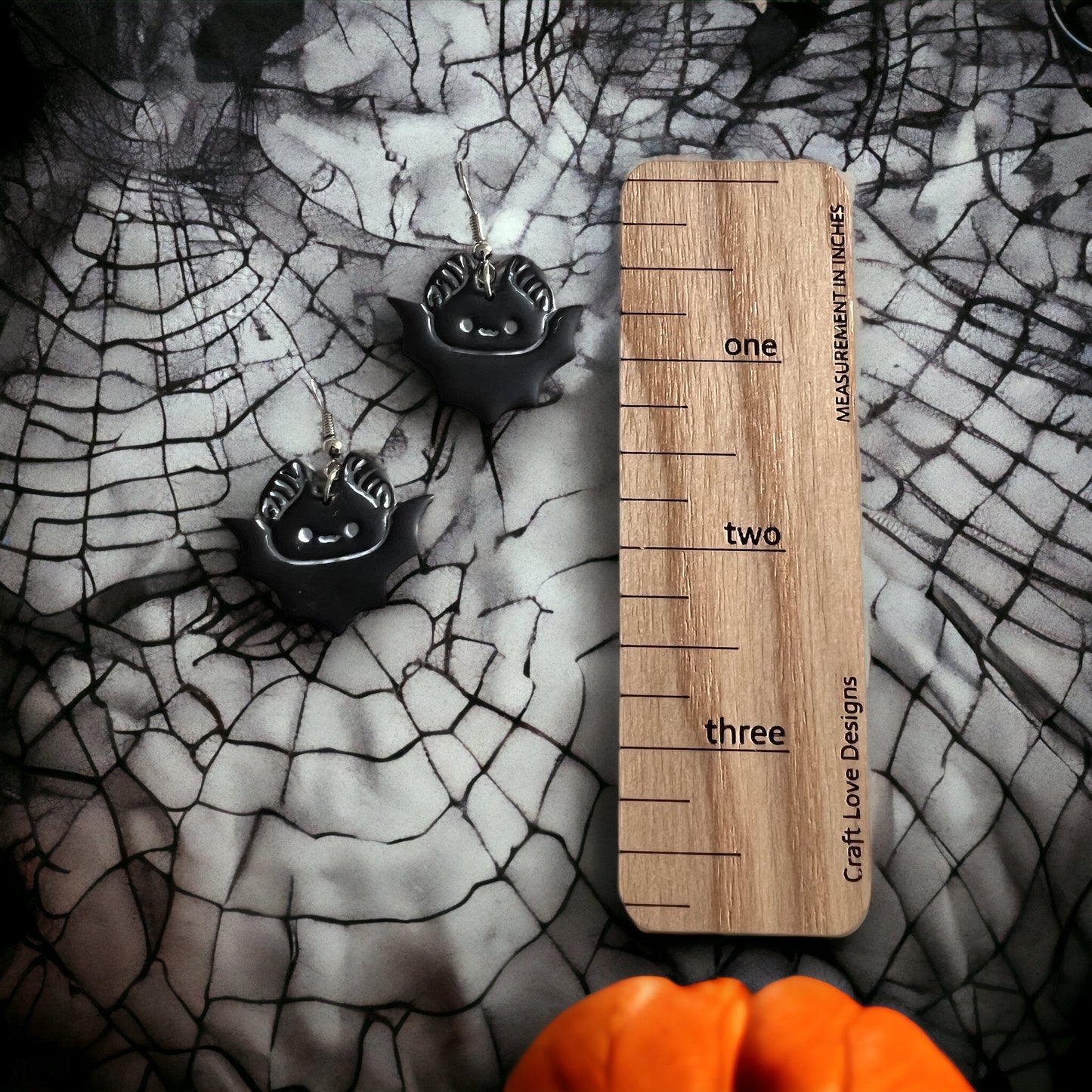 Close-up of bat dangle earrings resting on a spider-web-patterned background, next to an orange pumpkin and a wooden measurement ruler.