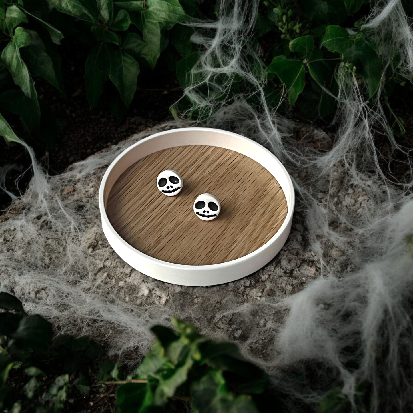 Round Jack Skellington-themed earrings resting on a wooden tray placed on a stone surface, surrounded by eerie cobwebs and ivy for Halloween vibes.