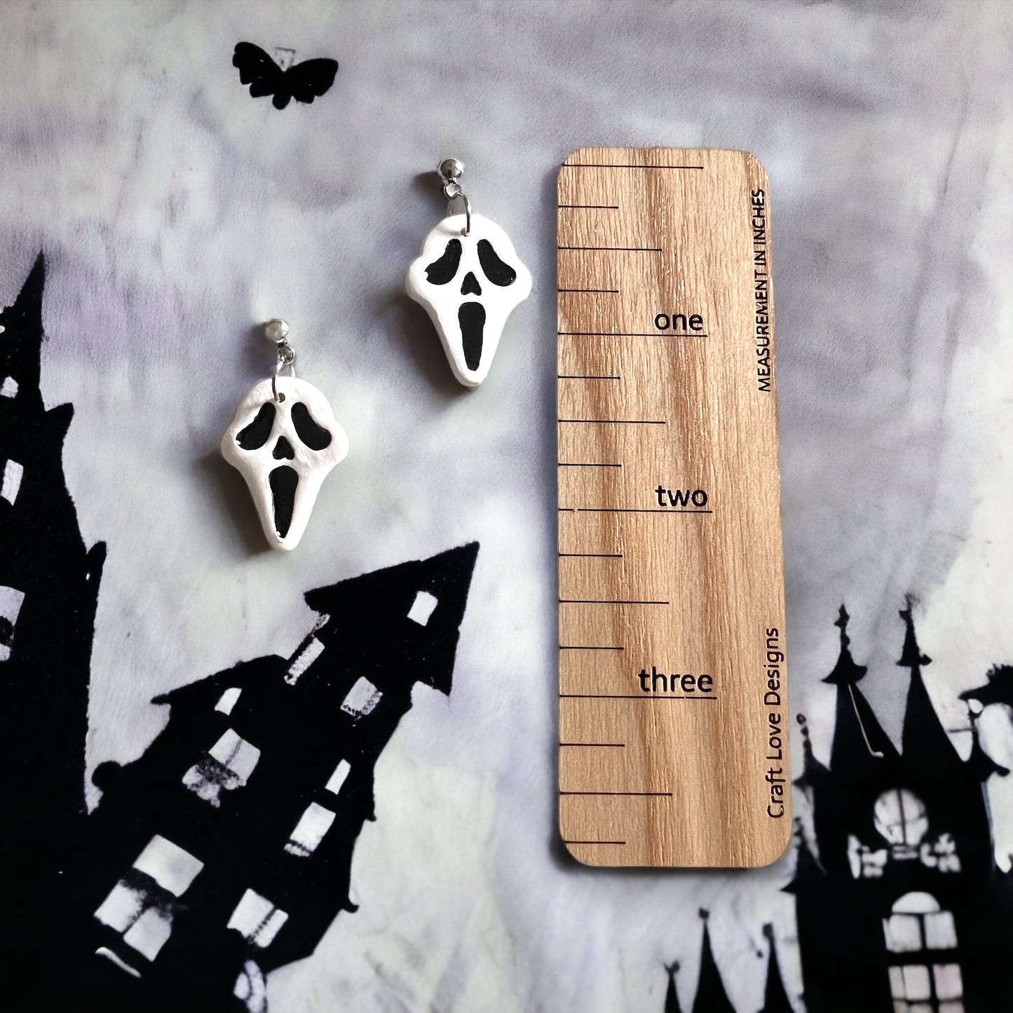White and black spooky face dangle earrings placed next to a wooden ruler, showcasing their size, with a Halloween-inspired background of haunted houses.