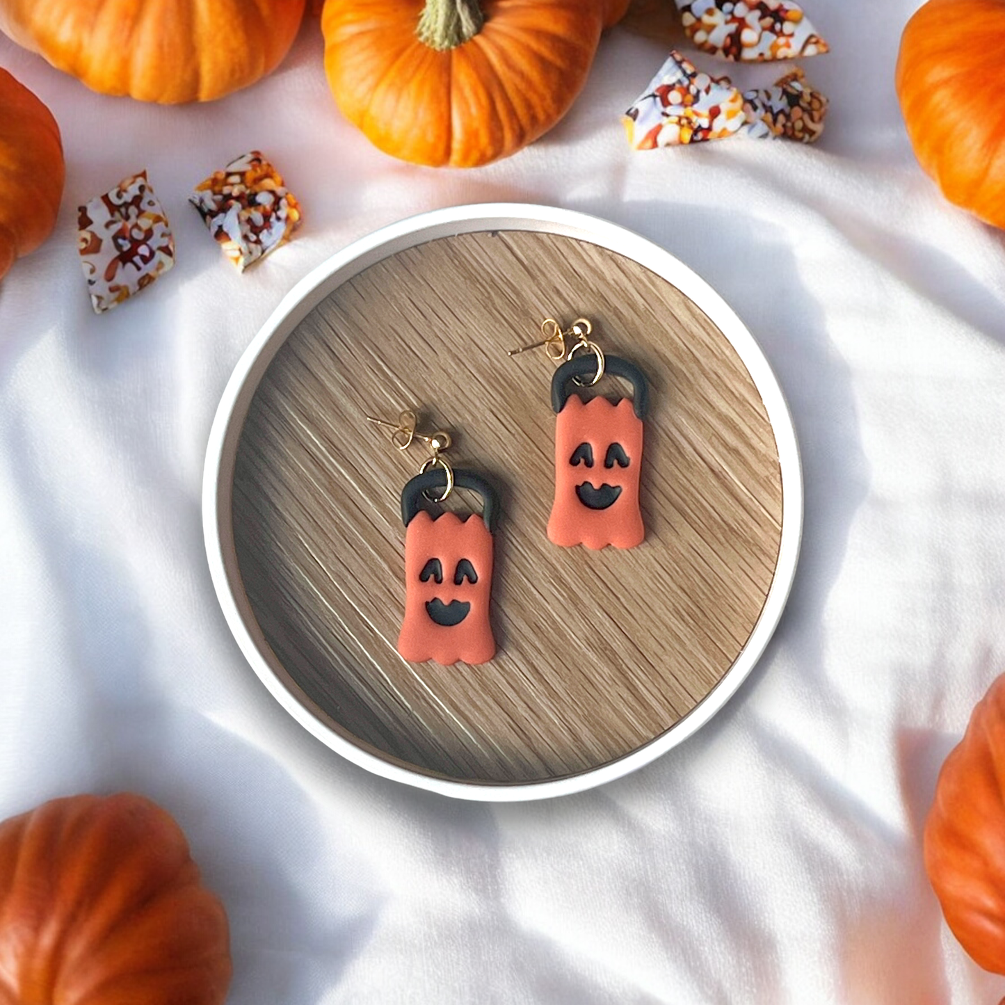 Pumpkin-shaped earrings with yellow "Spooky" text resting in a wooden tray, surrounded by red fabric and spooky Halloween accents.