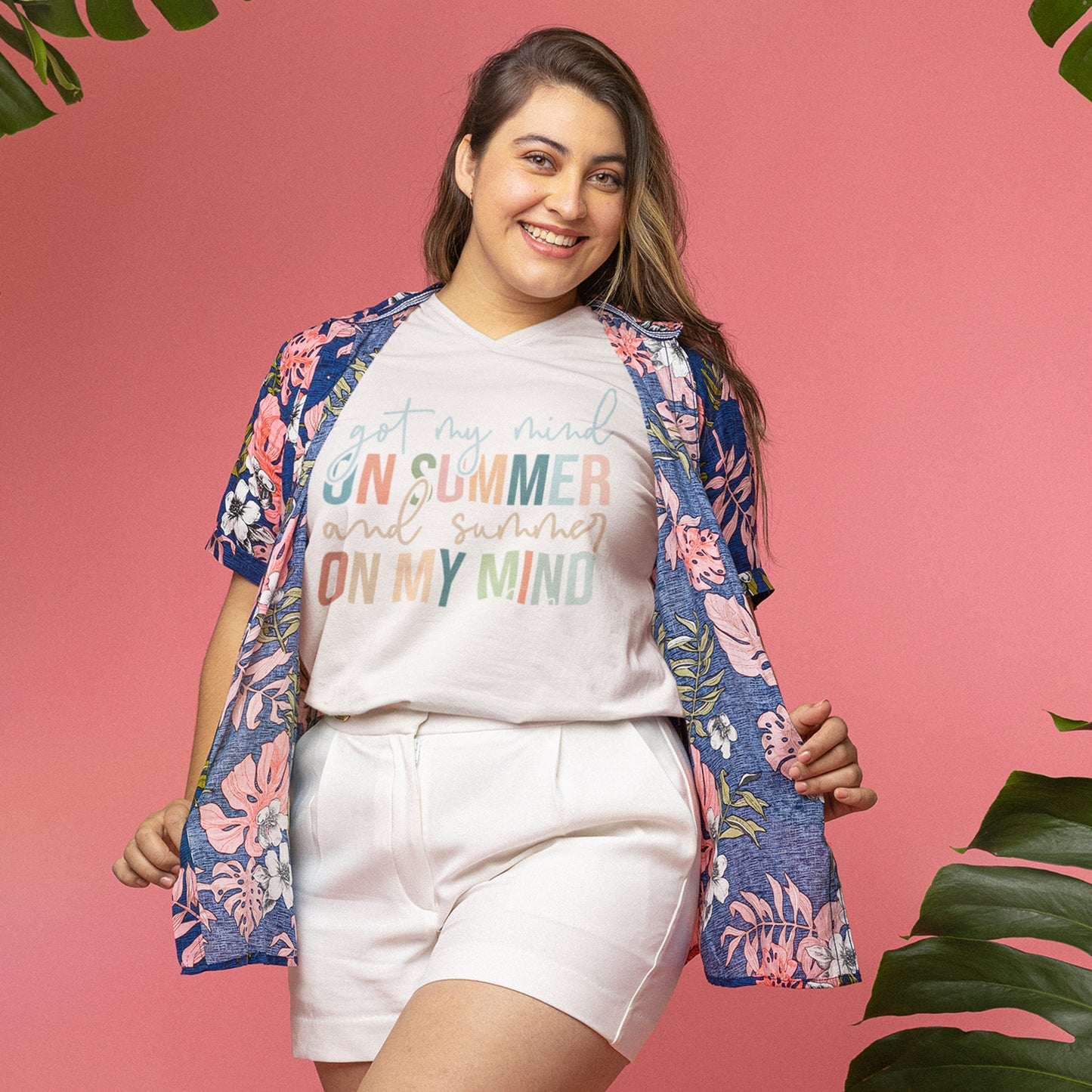 Woman wearing a summer quote t-shirt styled with a floral shirt and shorts, smiling against a pink background with tropical vibes.