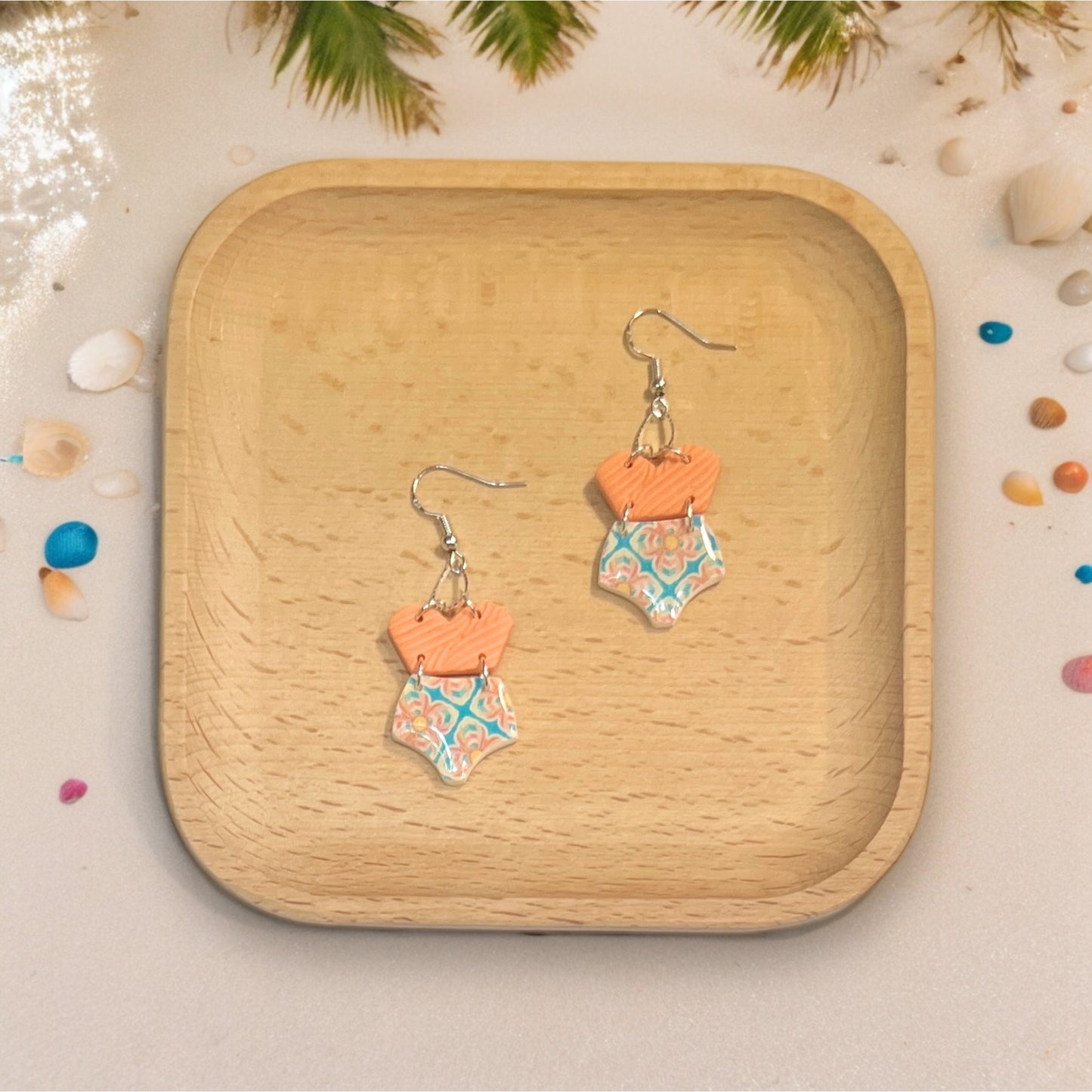 Fun polymer clay earrings with summer swimsuit designs placed on a wooden tray, surrounded by beach-themed accents.