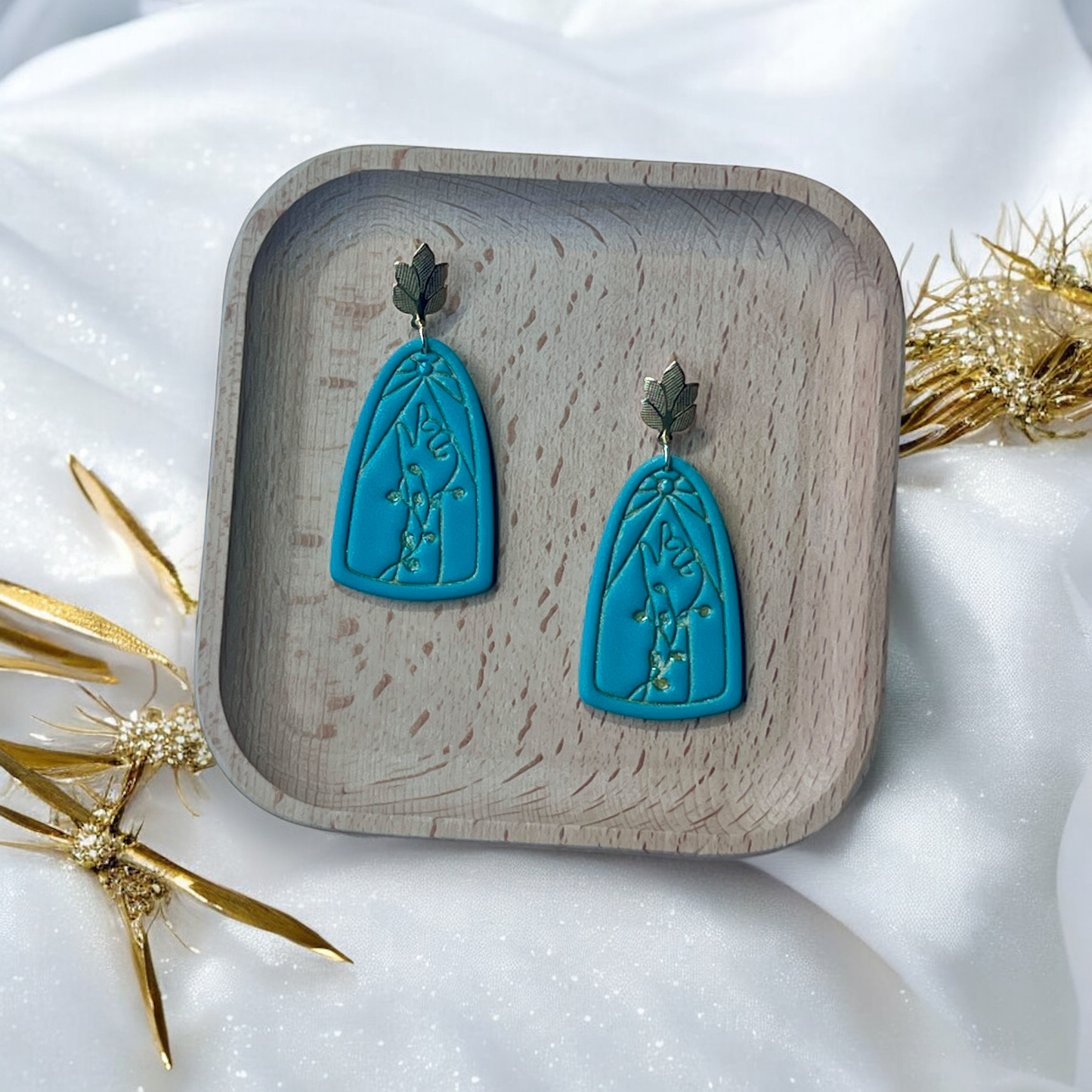 Teal arch-shaped earrings with gold leaf studs resting in a wooden tray, surrounded by white fabric and golden accents.