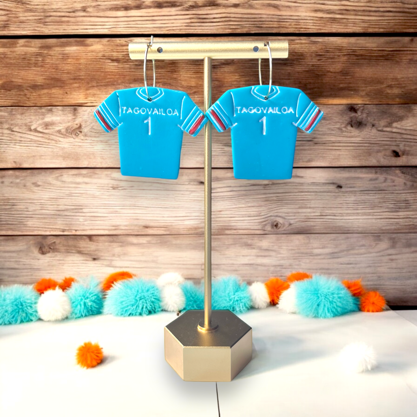 Teal Tua Tagovailoa sports jersey earrings on a decorative stand, surrounded by teal, orange, and white pom-pom accents