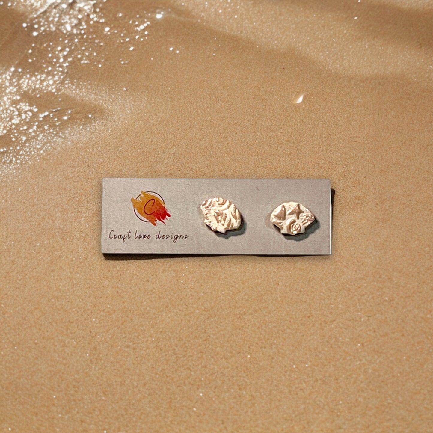 Pair of handmade textured earrings on a branded Craft Love Designs card, displayed on warm sand for a coastal-inspired aesthetic.