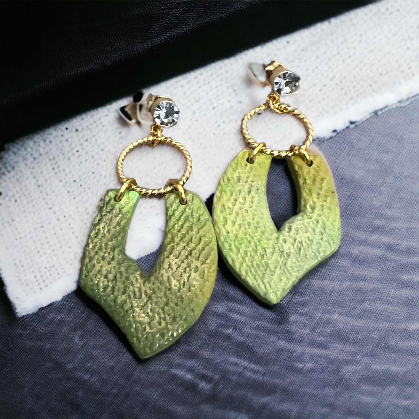 Close-up of textured green boho earrings featuring leaf-shaped pendants, gold detailing, and cubic zirconia studs on a fabric background.