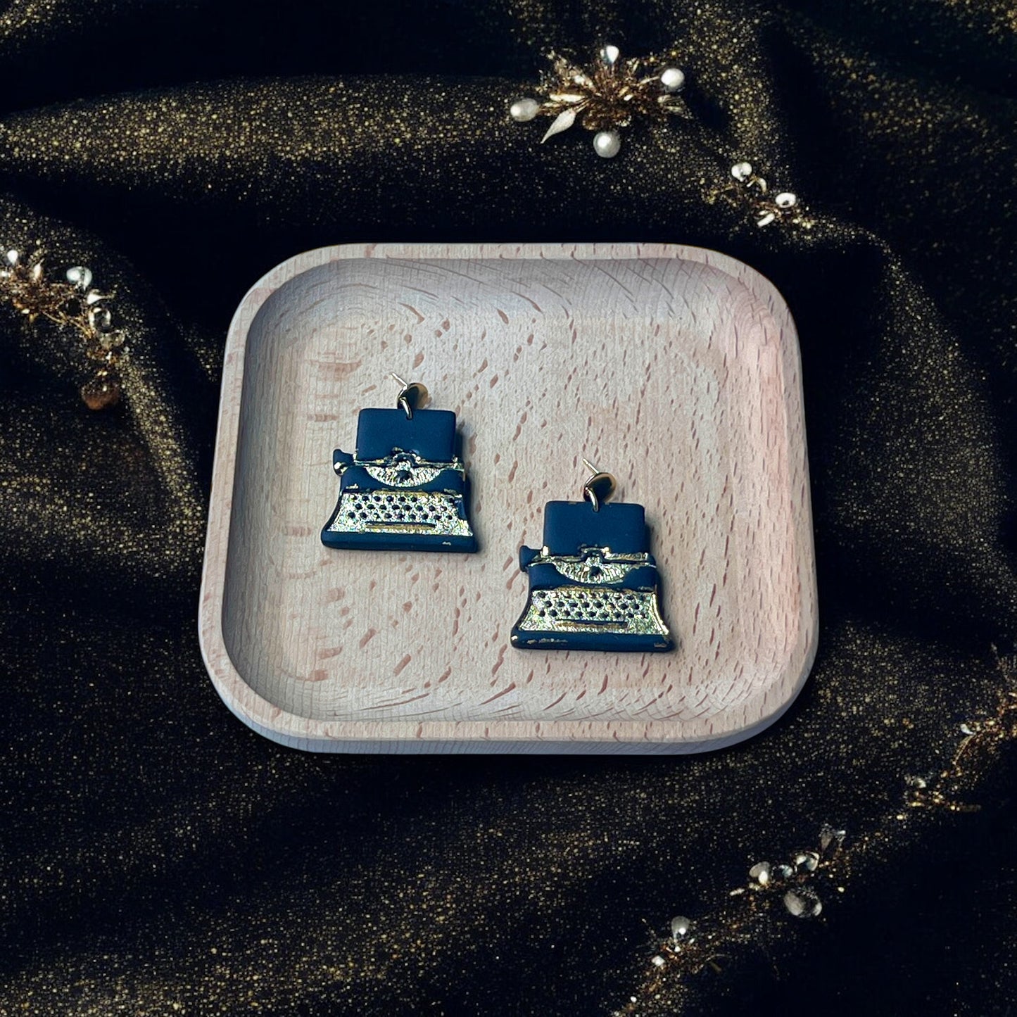 Vintage-style black and gold typewriter earrings resting in a wooden tray, surrounded by sparkly fabric, pearls, and gold accents.