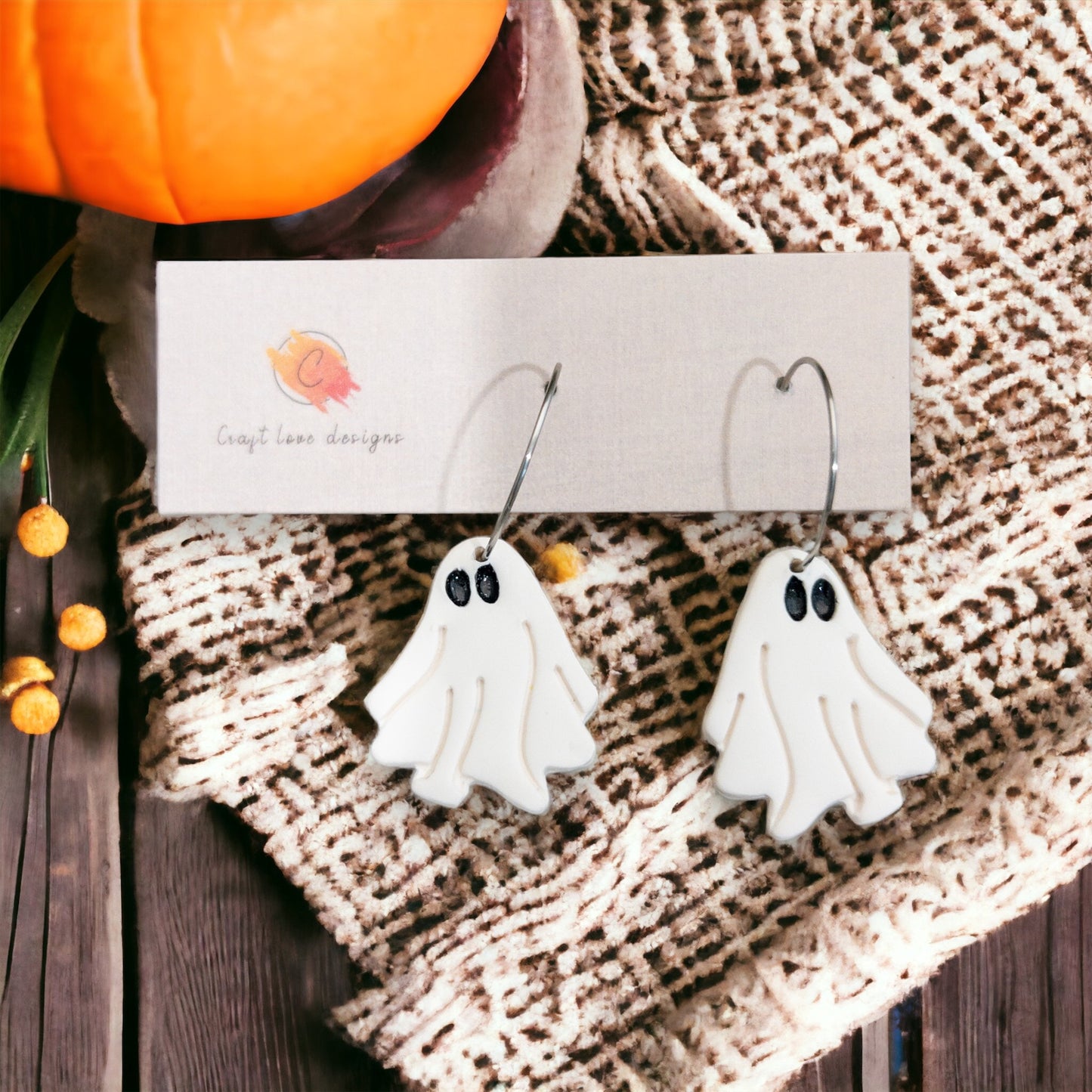 Pair of white spooky ghost-shaped dangle earrings resting on a cozy autumn-themed background with a pumpkin nearby.
