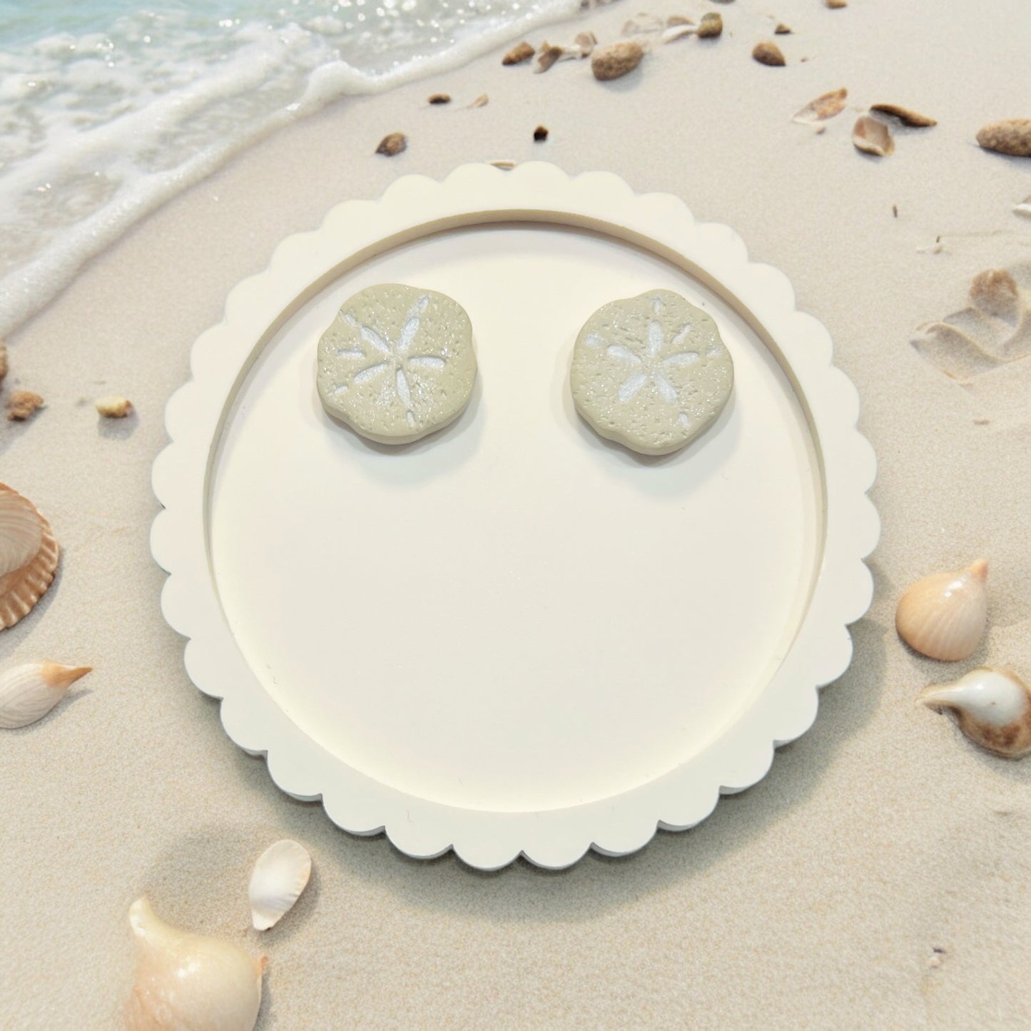 White handcrafted sand dollar earrings on a round decorative plate set against a beach backdrop, surrounded by seashells.