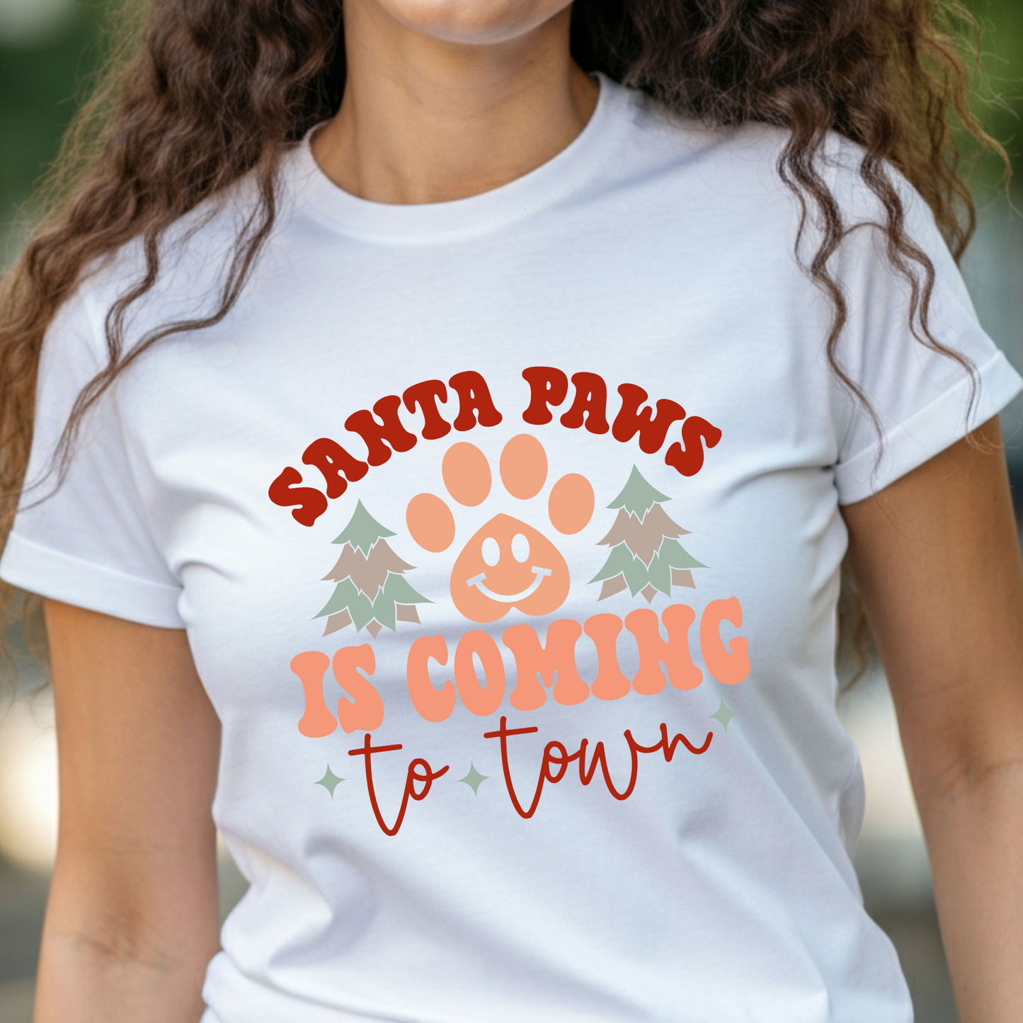 Woman wearing a white t-shirt with a festive "Santa Paws Is Coming to Town" design, complete with paw print and holiday graphics.