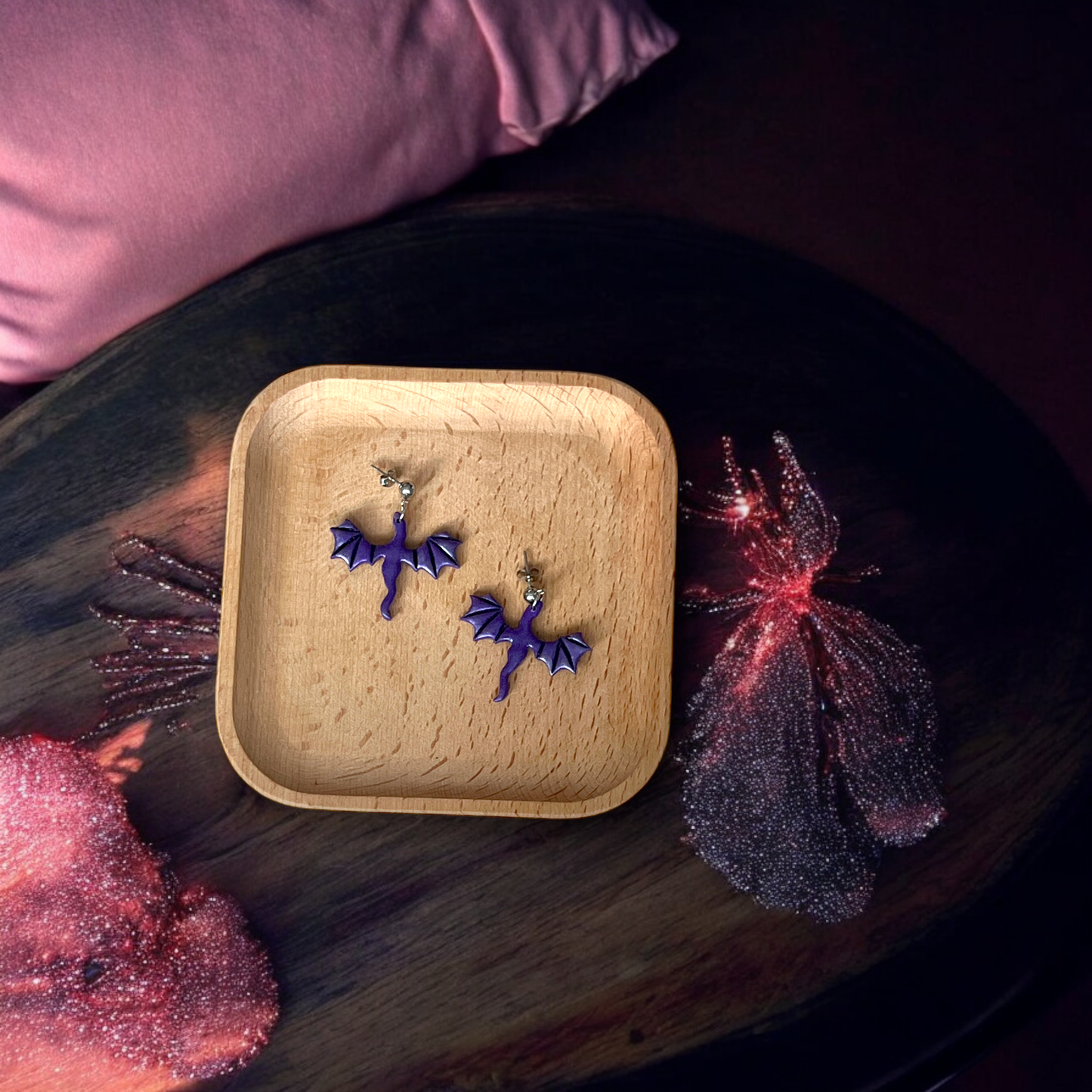 Purple bat earrings resting in a wooden tray, surrounded by sparkly pink decorations and soft fabric in a dark, mystical setting.