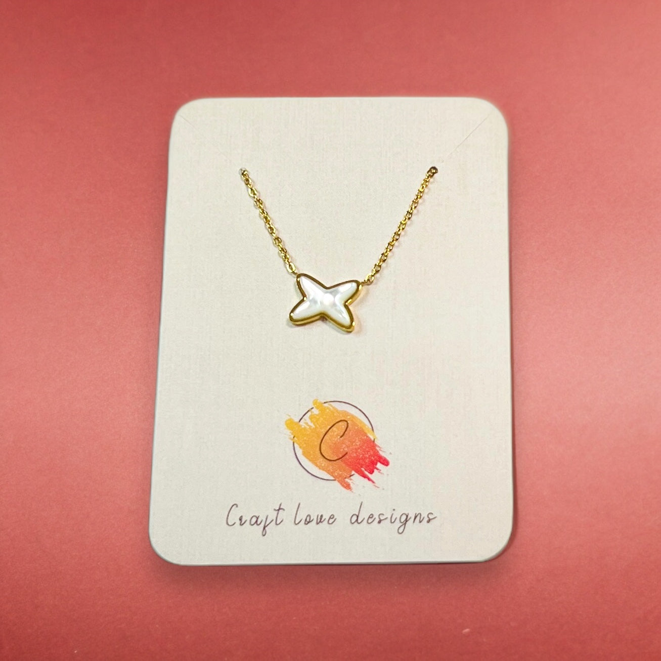 Gold necklace with a pearly X shell pendant displayed on a Craft Love Designs card against a coral backdrop, perfect for everyday wear.