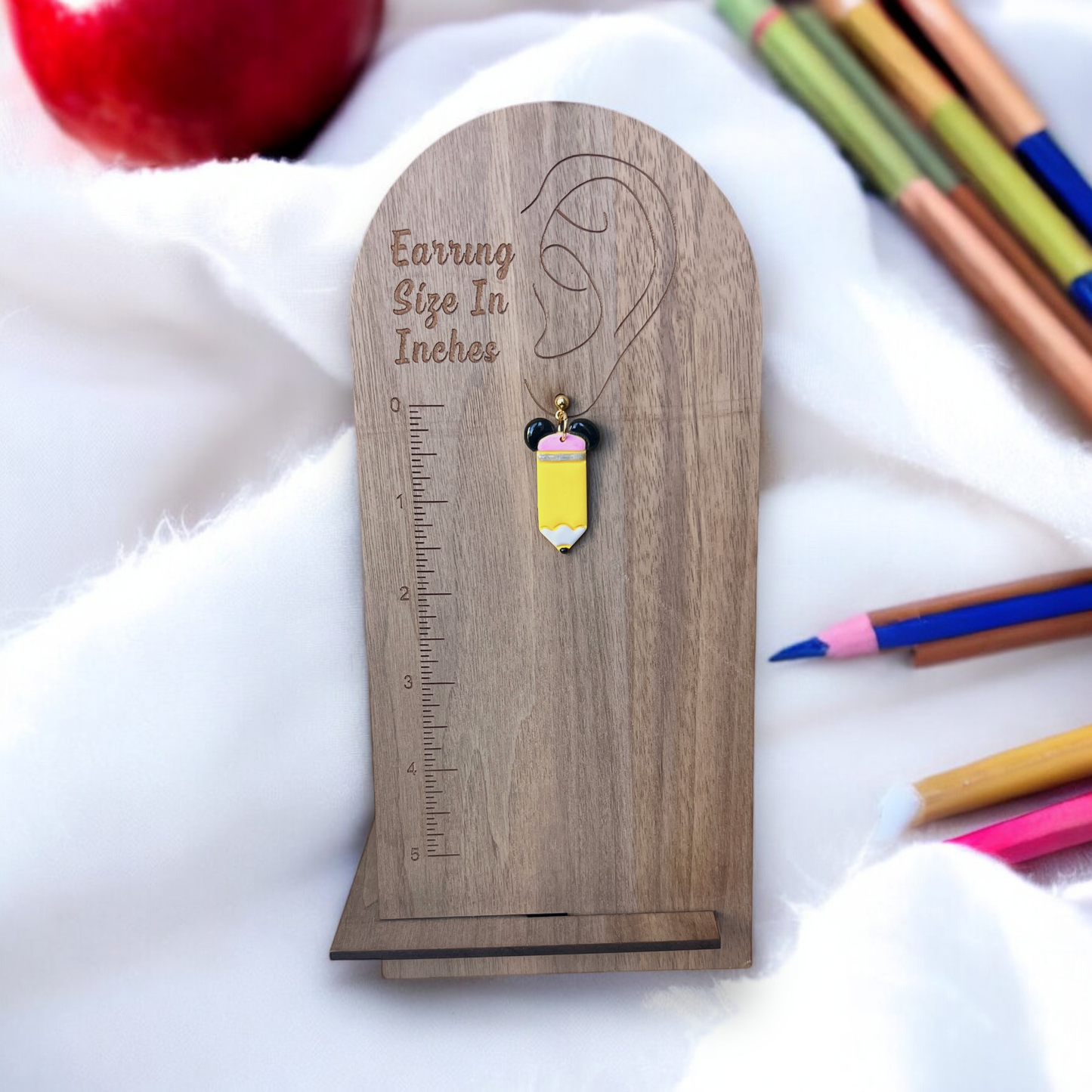 Single yellow pencil-themed earring with black ears displayed on a wooden size guide stand, placed over a soft fabric with colorful pencils nearby.