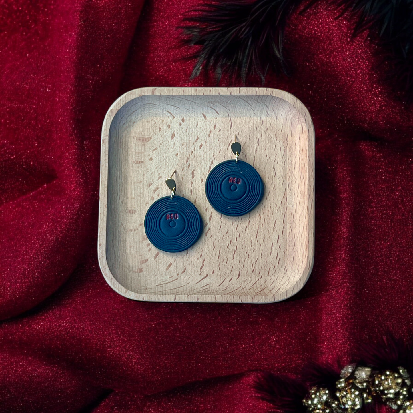 Retro vinyl record-style earrings with black grooves and "Red" detailing, placed in a wooden tray with a rich red velvet fabric background.