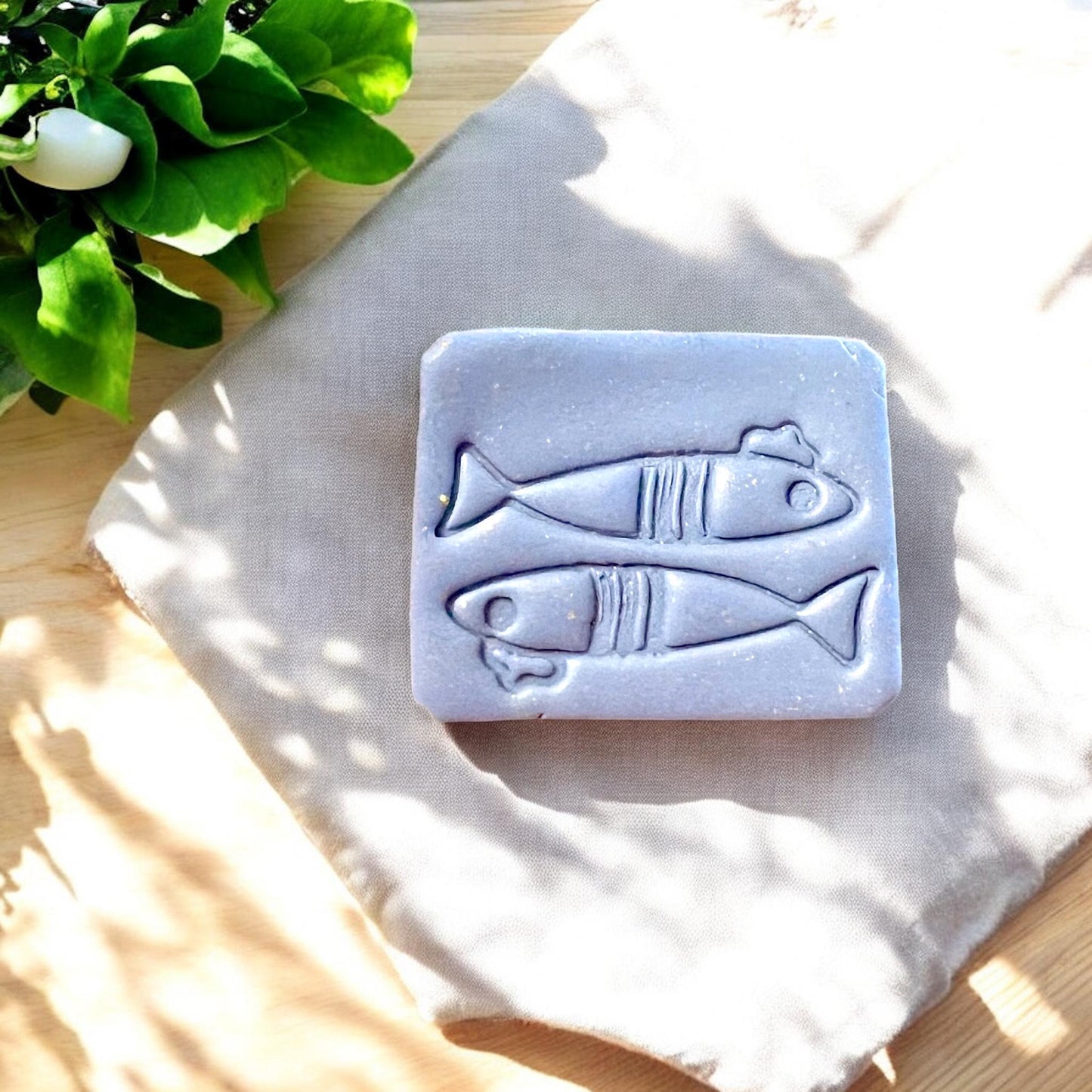 Two clay fish charms with etched details and French hats, styled on soft neutral linen with leafy greens and sunlight shadows
