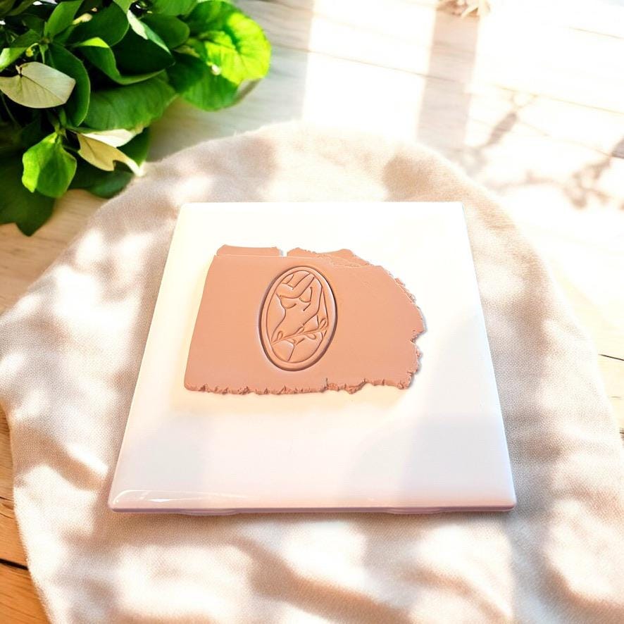 Oval female silhouette cutter impressed in nude-toned clay on ceramic tile with soft fabric backdrop.
