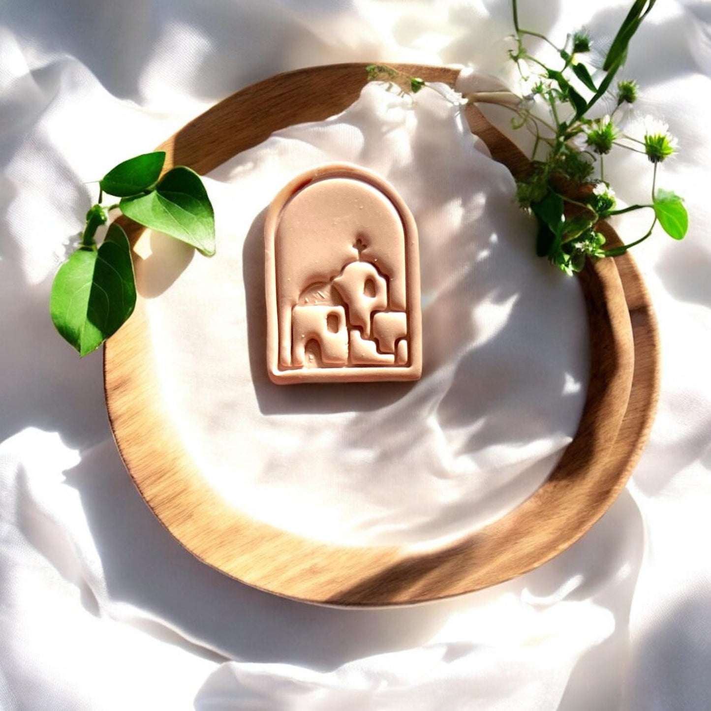Close-up of finished clay earring piece with Santorini village scene, placed on a wooden round tray with white fabric and green leaves.